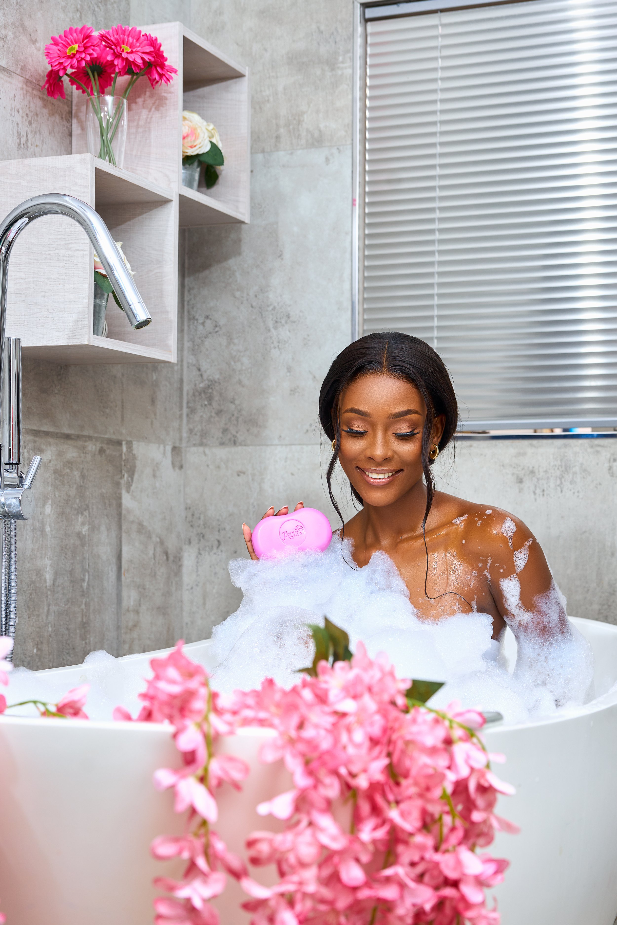 A smiling woman with dark hair in a bathtub filled with bubbles, holding a pink bath bomb, surrounded by pink flowers with a background of a modern bathroom with a window and shelves with flowers.