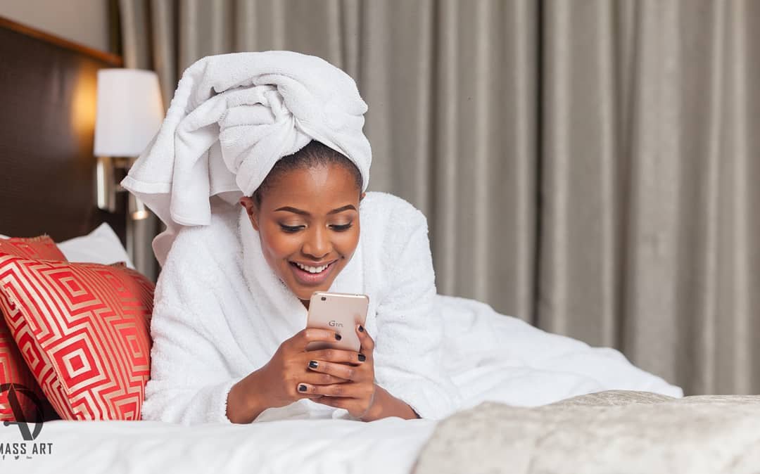 A woman with a towel wrapped on her head, lying on a bed, smiling and looking at her phone in a hotel room.