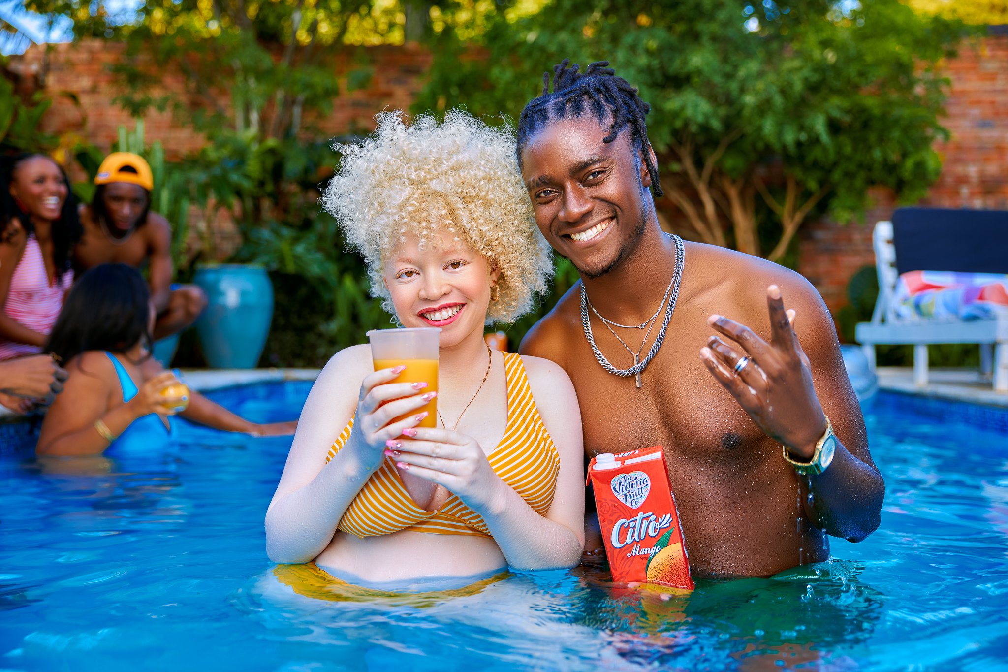 Group of young adults having fun in a backyard pool during daylight, with two people smiling at the camera in the foreground and others in the background enjoying drinks and conversation.