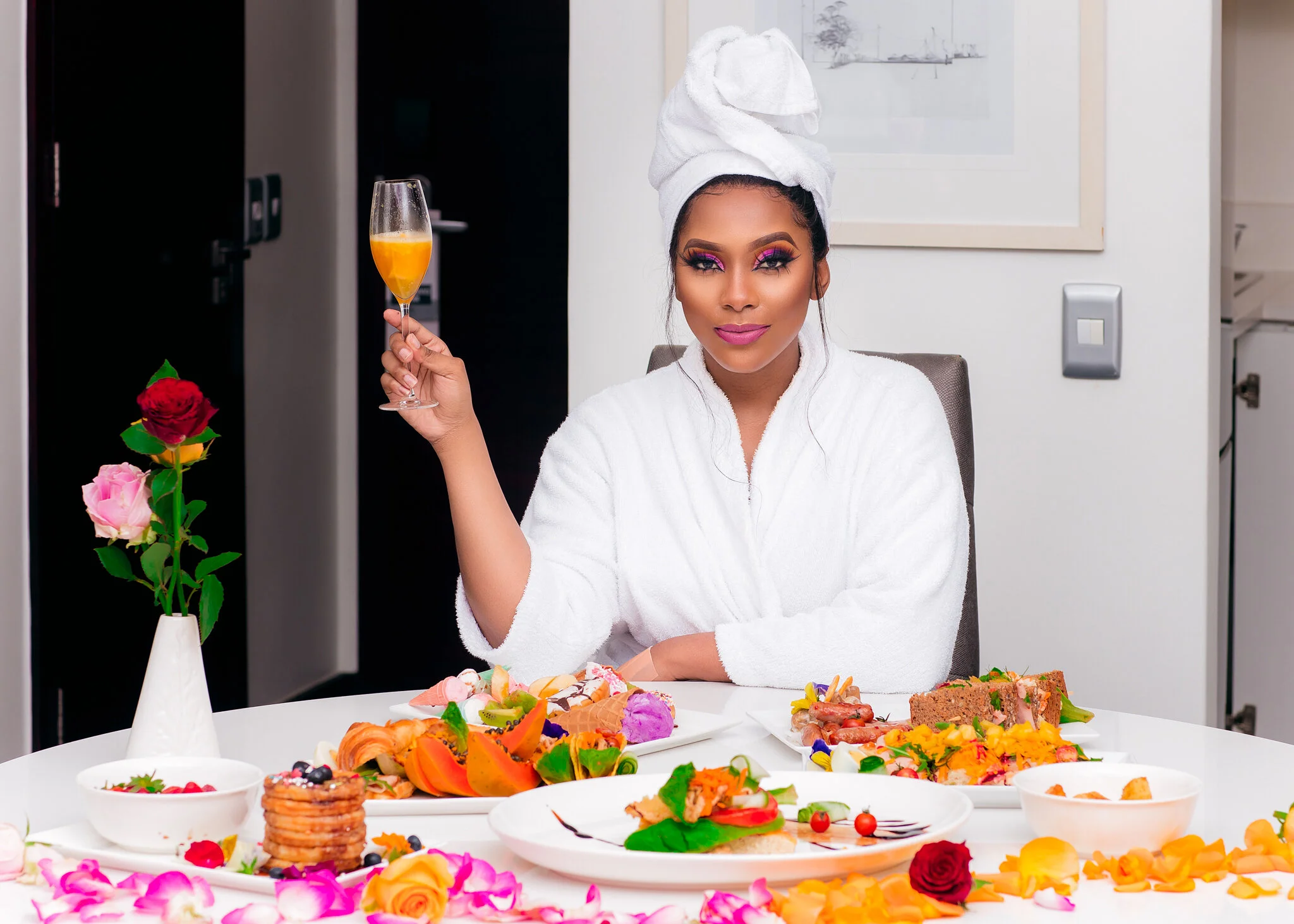 A woman in a white bathrobe with a towel wrapped around her head, holding a glass of orange drink, sitting at a table with various colorful foods and flowers.