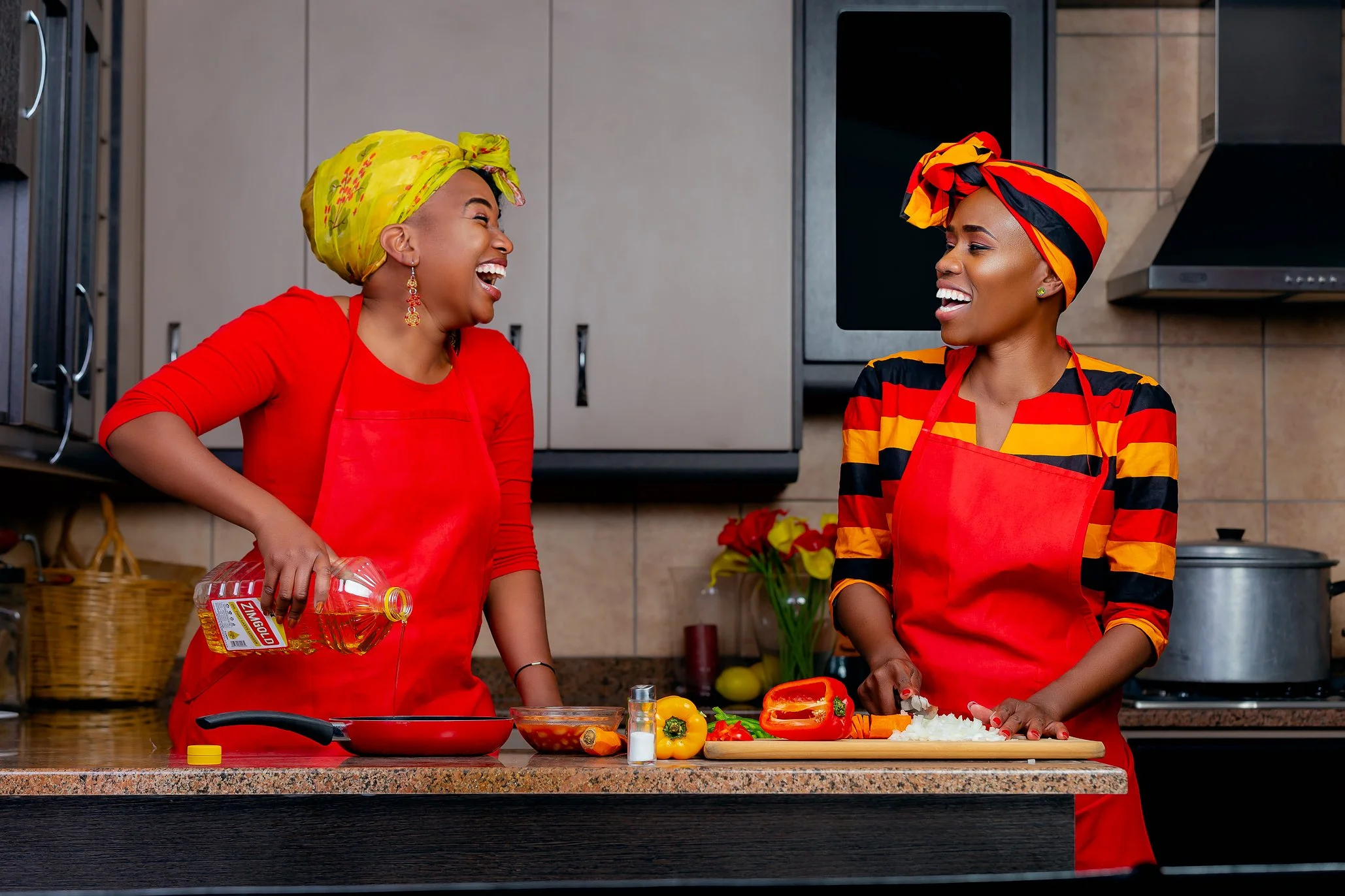 Two women in colorful head wraps and red aprons sharing a laugh in a kitchen while preparing vegetables for a meal.