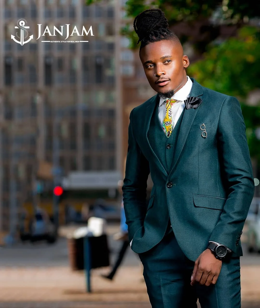 A young man in a dark suit with a white shirt, patterned tie, and black bow tie, standing outdoors with a cityscape background, posing confidently.