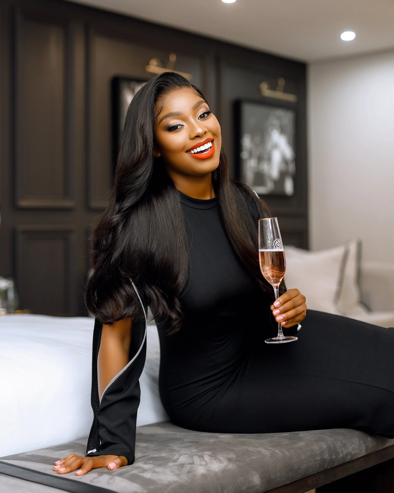 A woman with long, black, wavy hair, wearing a black dress and holding a glass of rosé champagne, sitting on a bed in a hotel room, smiling.