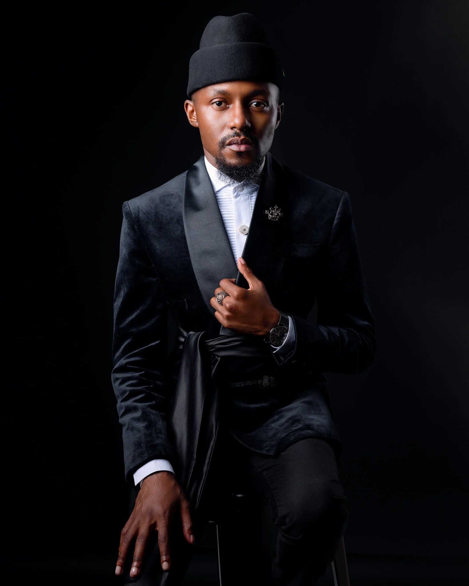 Portrait of a man wearing a black beanie, black blazer with satin lapel, white shirt, and accessories, seated against a dark background.