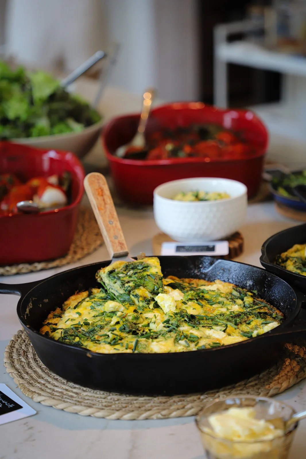 A cooked frittata with greens on a woven placemat, surrounded by other dishes and bowls of food on a dining table.