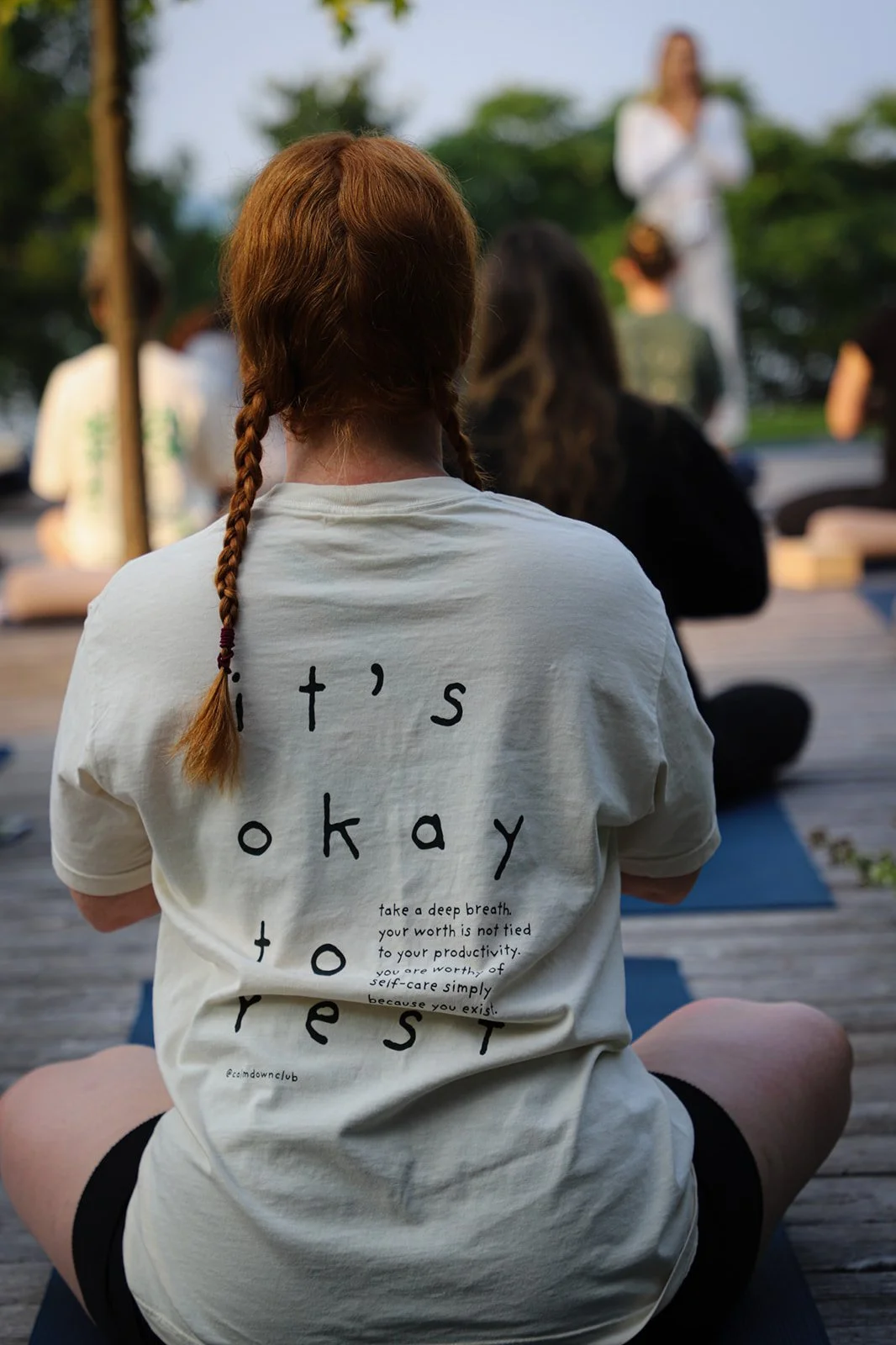 Person with red braided hair sitting on a yoga mat attending an outdoor meditation or yoga class, with a woman speaking at the front in the background.
