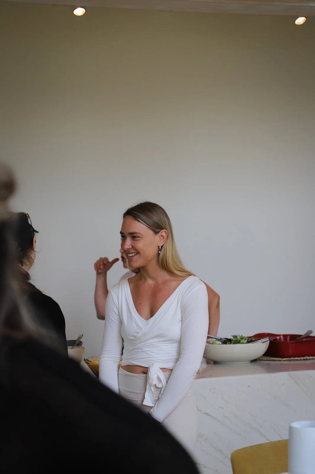 Woman with blonde hair smiling and talking to another person in a kitchen or dining area.