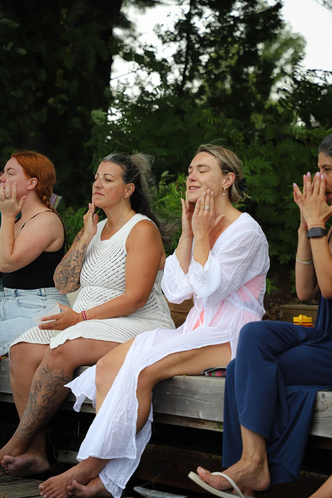 Four women sitting outdoors with their eyes closed and hands on their faces, possibly meditating or participating in a calming activity, against a background of large green trees.