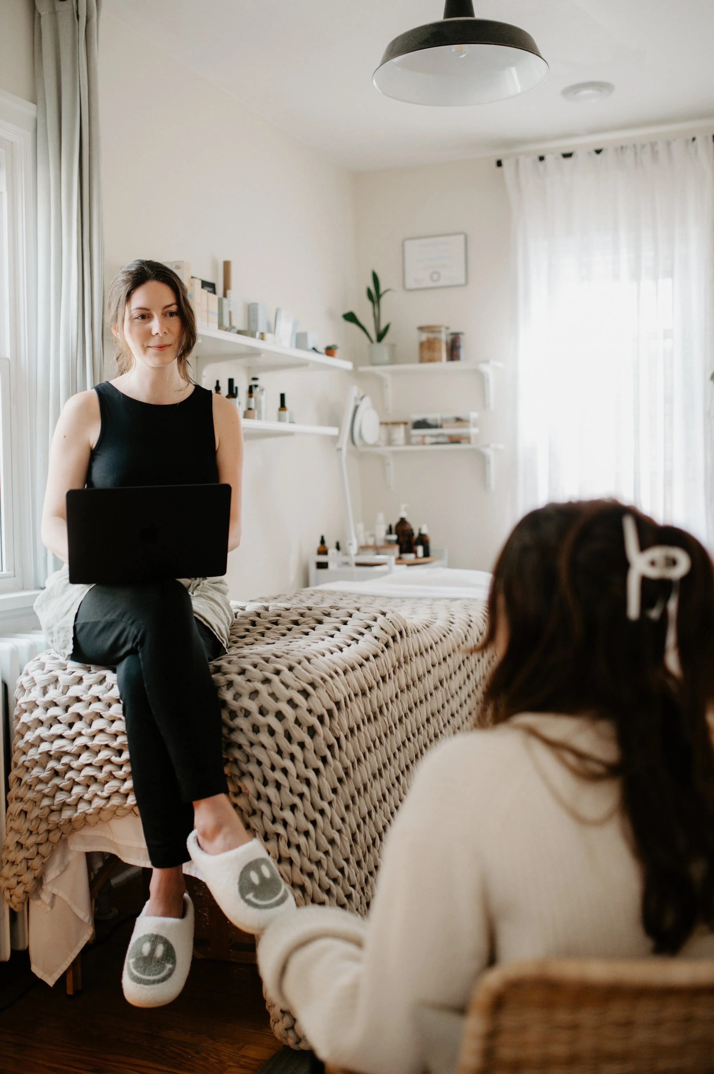 A woman sitting on a massage table with a laptop, talking to a woman sitting nearby in a cozy, well-lit room, possibly a therapy or massage room.