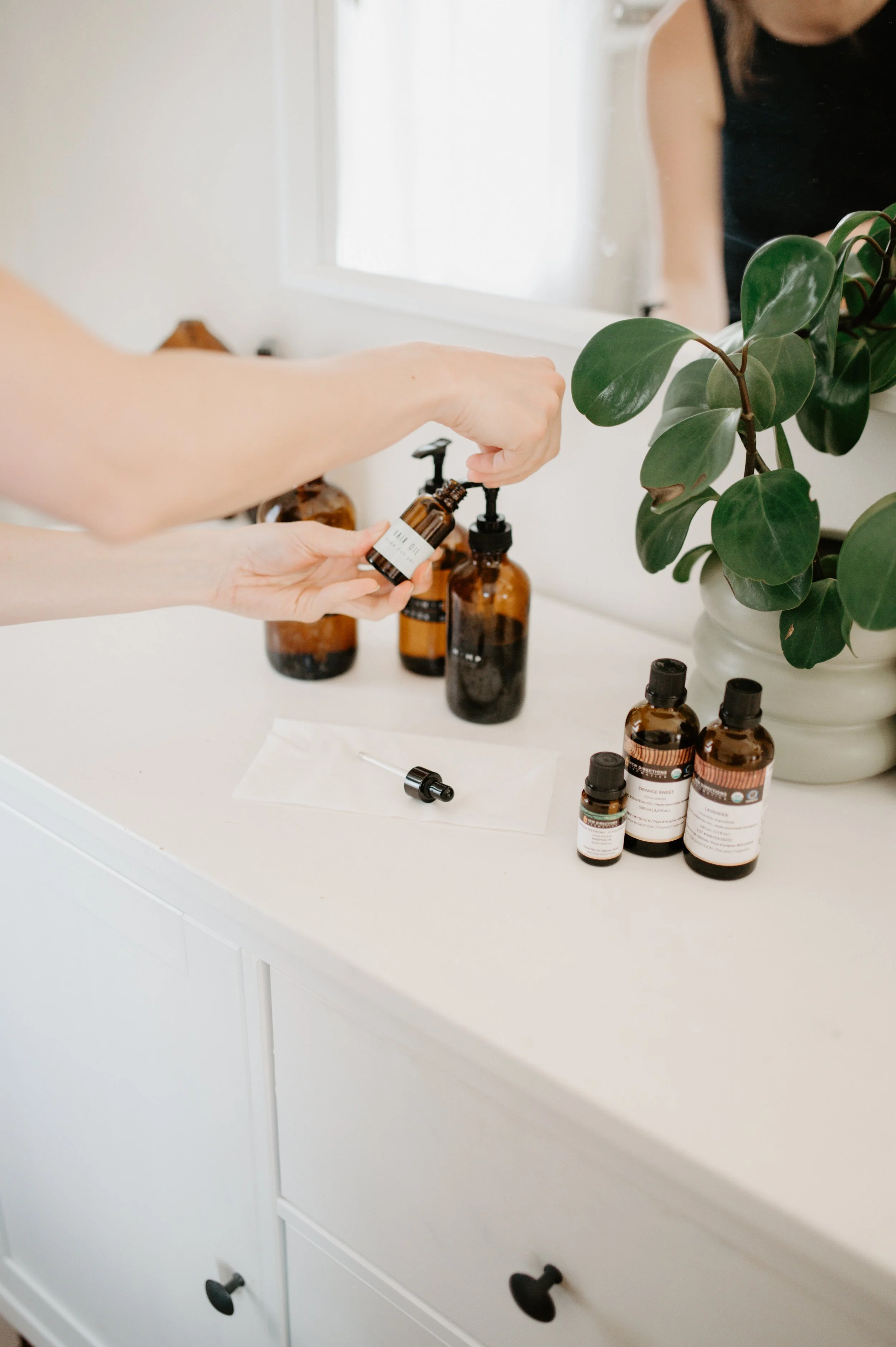 Person pouring essential oils from amber bottles onto a white surface, with a large green plant in a white pot on a white dresser.