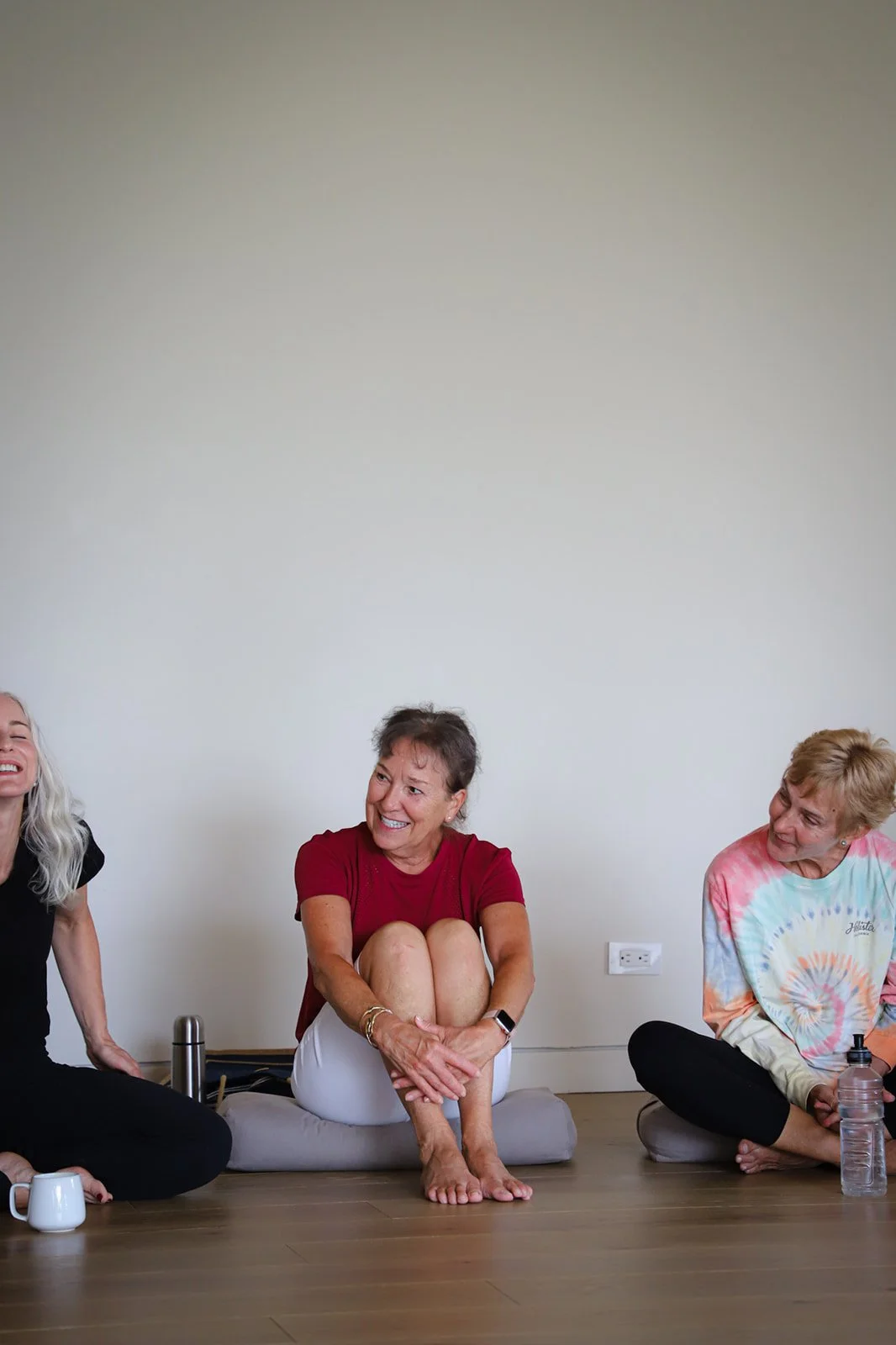 Three women sitting on floor cushions, laughing and talking during a casual gathering in a room with plain wall and hardwood floor.