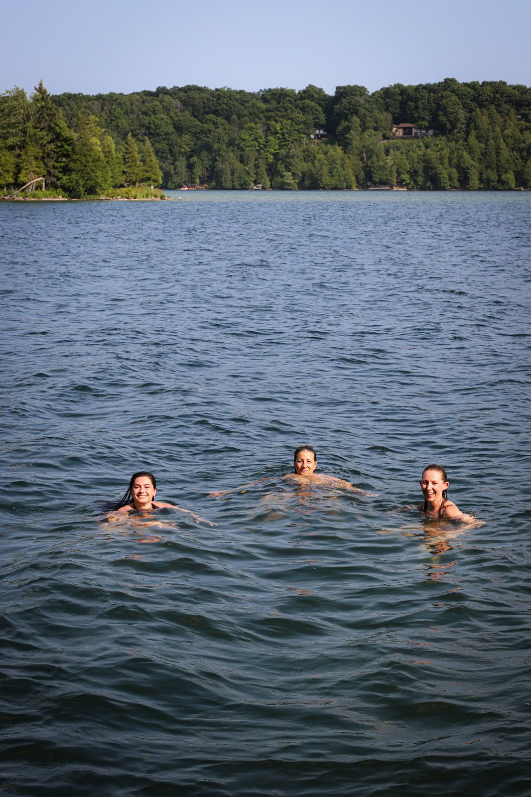 Three young women swimming in a large lake surrounded by green trees and a clear sky.