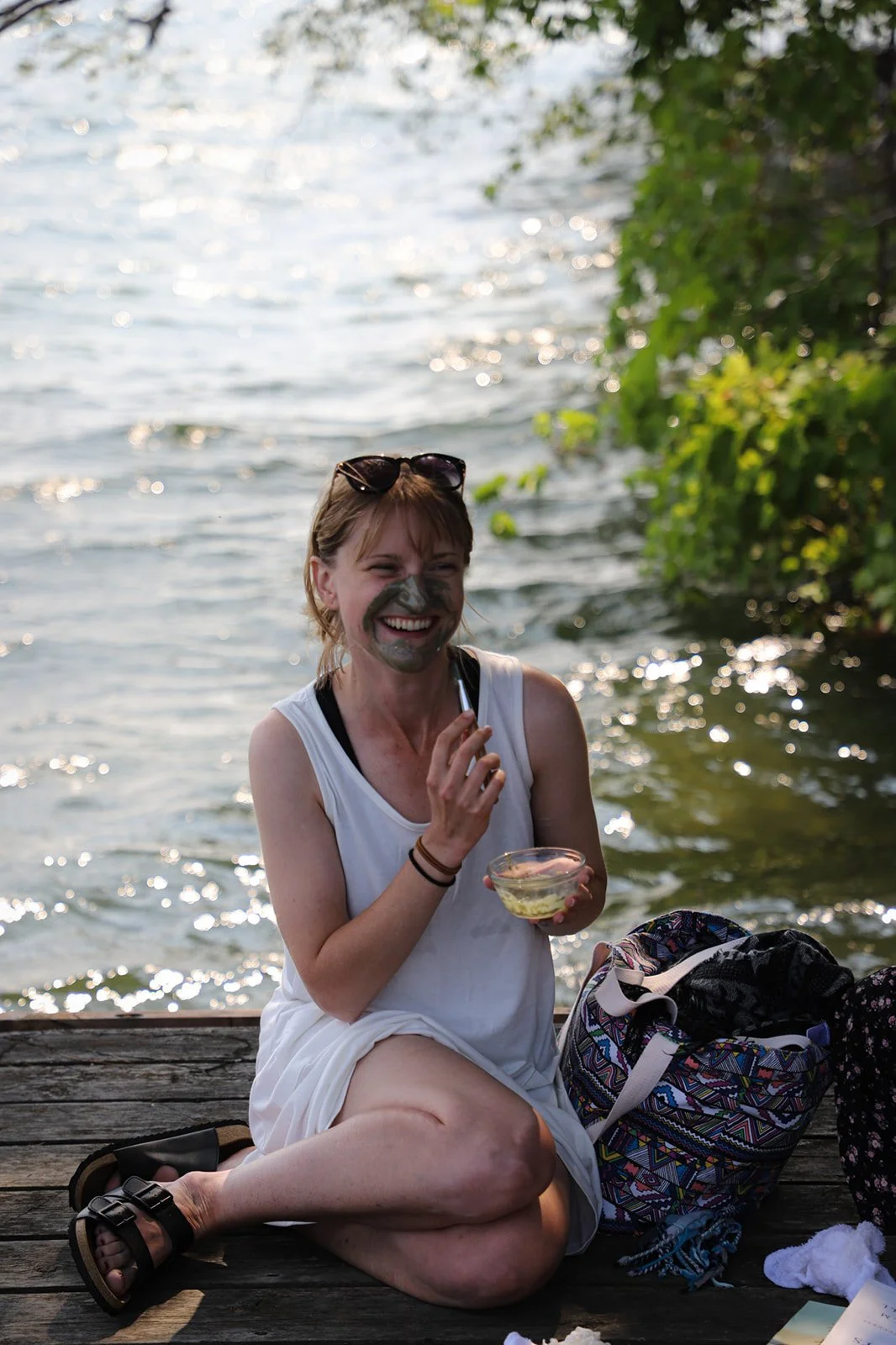 A woman sitting on a dock near a body of water, smiling and applying mud face mask, with a colorful bag, towel, and other personal items nearby.