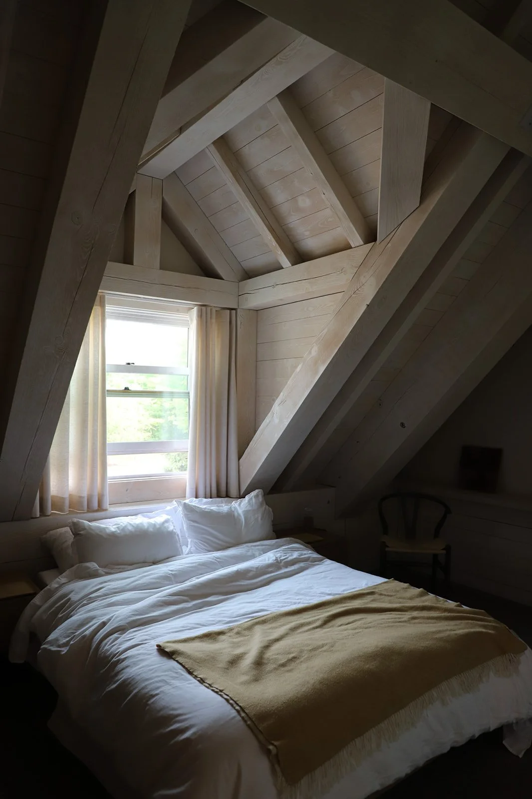 A cozy bedroom with a wooden ceiling and walls, a window with white curtains, a bed with white bedding and pillows, and a yellow blanket, in a room with sloped walls.