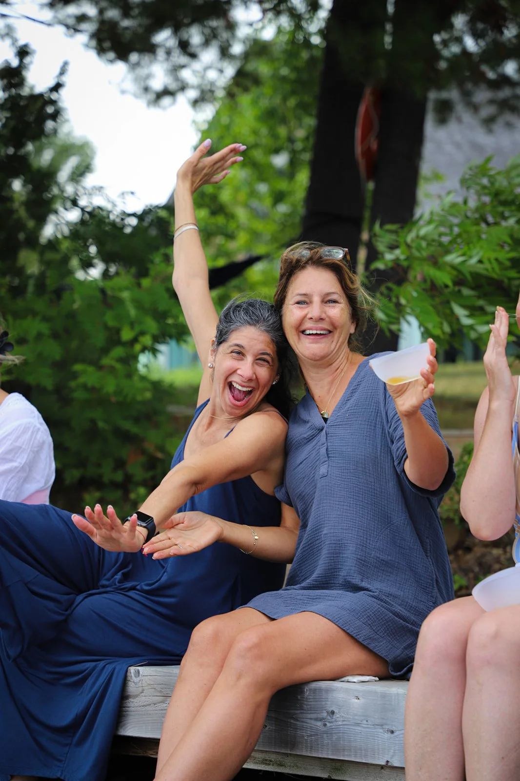 Two women sitting on a wooden bench outdoors, smiling and enjoying a happy moment together. One woman has long dark hair and the other has short light brown hair, and they are surrounded by green trees.