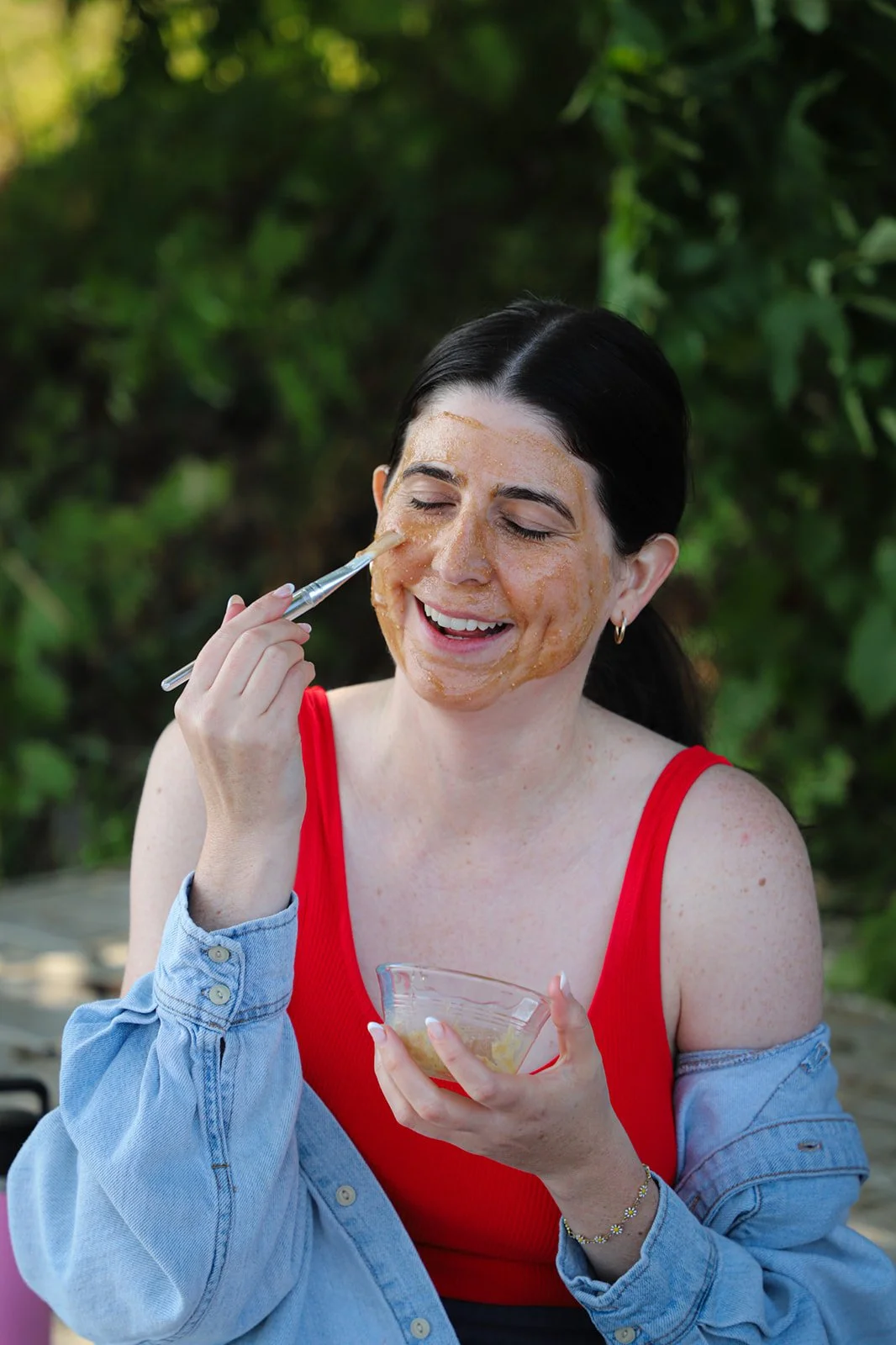 A woman in a red top and denim jacket applying a facial mask outdoors, smiling with eyes closed, holding a bowl of the mask, with green foliage background.