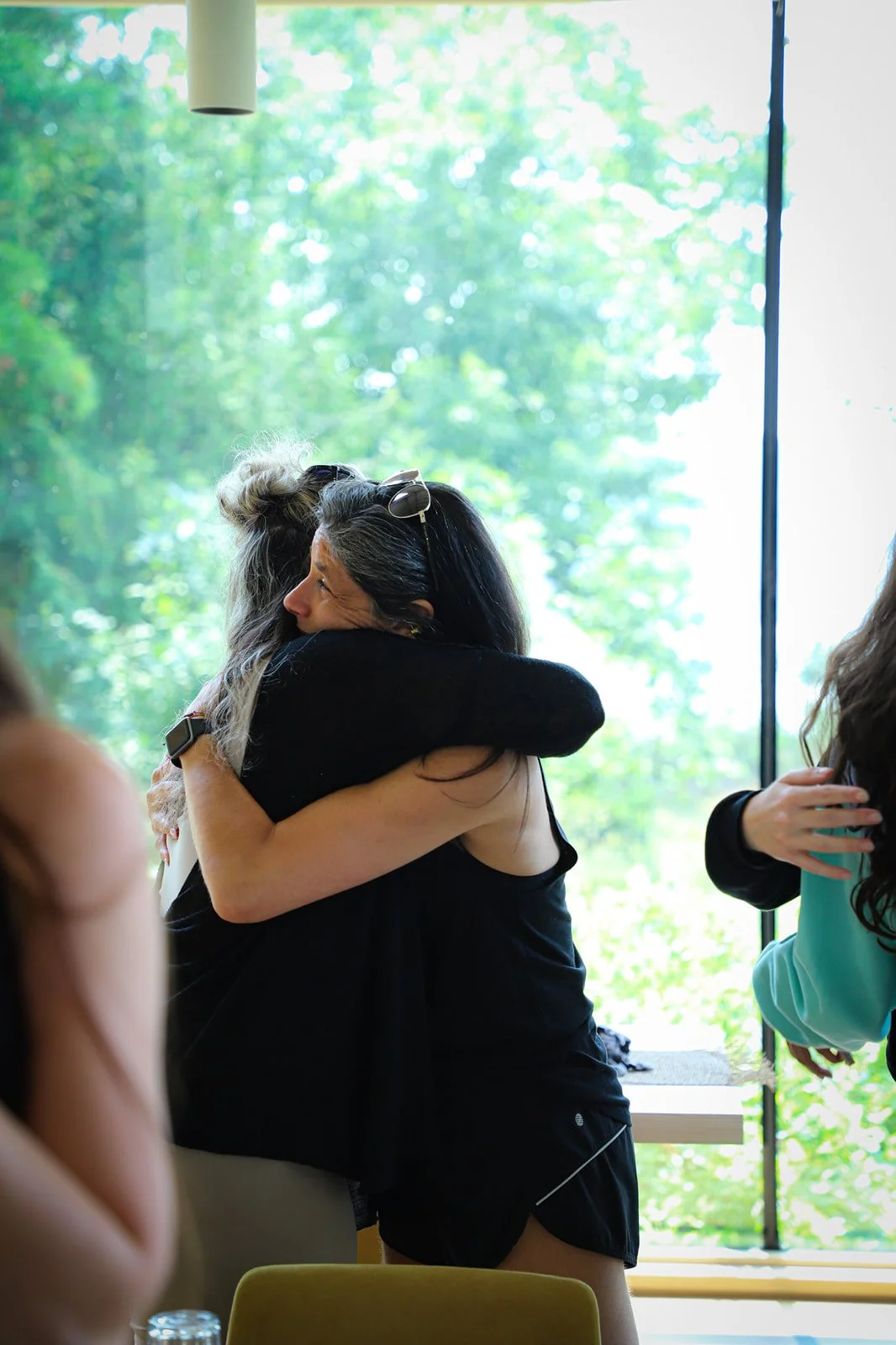 Two women hugging each other warmly inside a building with large windows showing green trees outside.