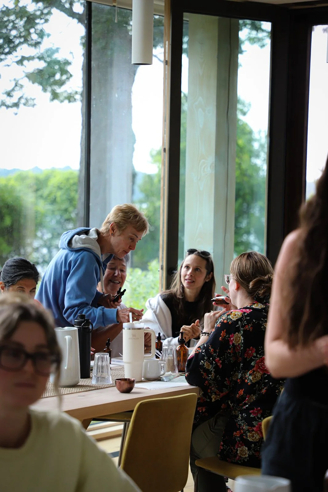 A group of people gathered around a table having a conversation inside a room with large windows showing greenery outside.