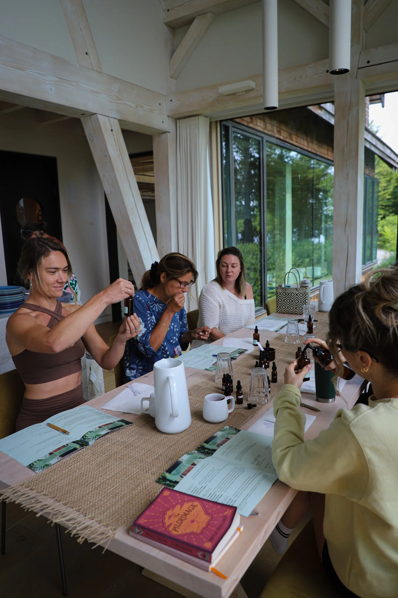 Group of women sitting at a wooden table, examining essential oils, with bottles and syrup on the table, in a modern room with large windows and natural light.