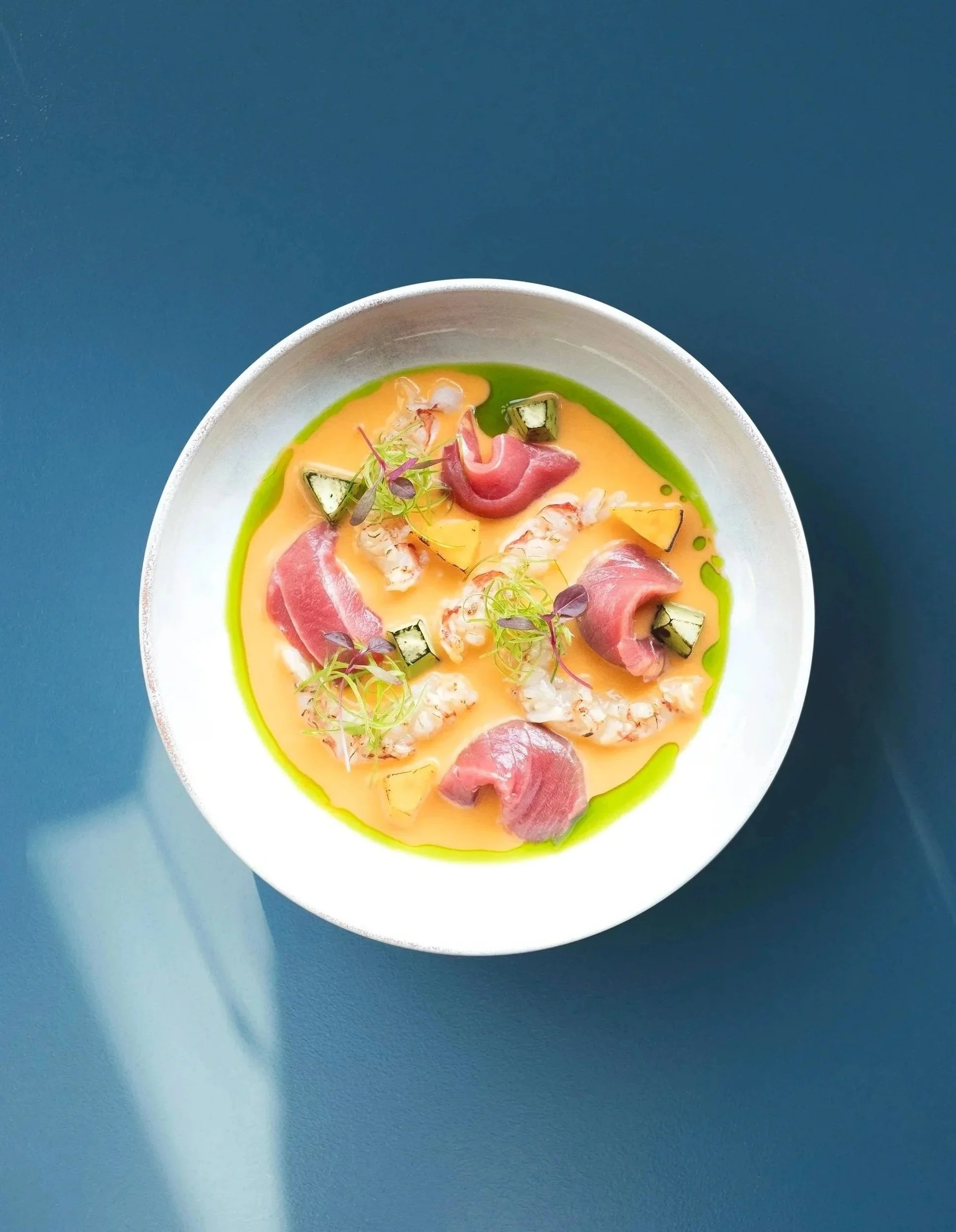 A white bowl containing a dish with pink slices of meat, green sauce around the edges, small chopped vegetables, and microgreens, set on a dark teal surface.
