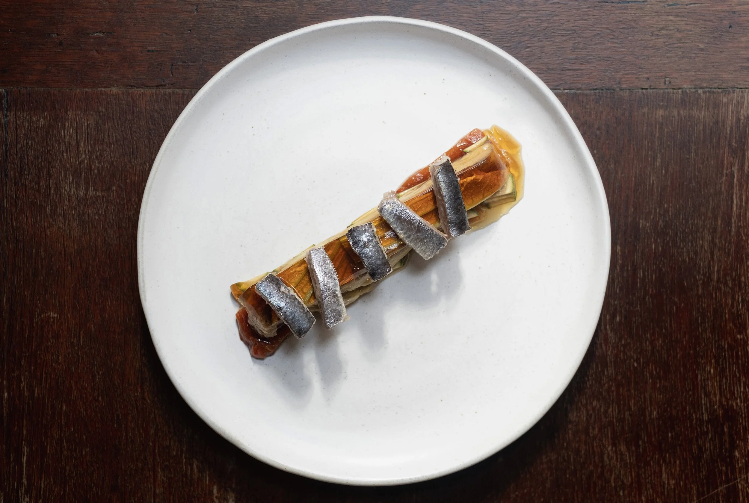 A plated dish featuring a cylindrical roll with vegetables and topped with sardine fillets on a white plate, placed on a wooden surface.
