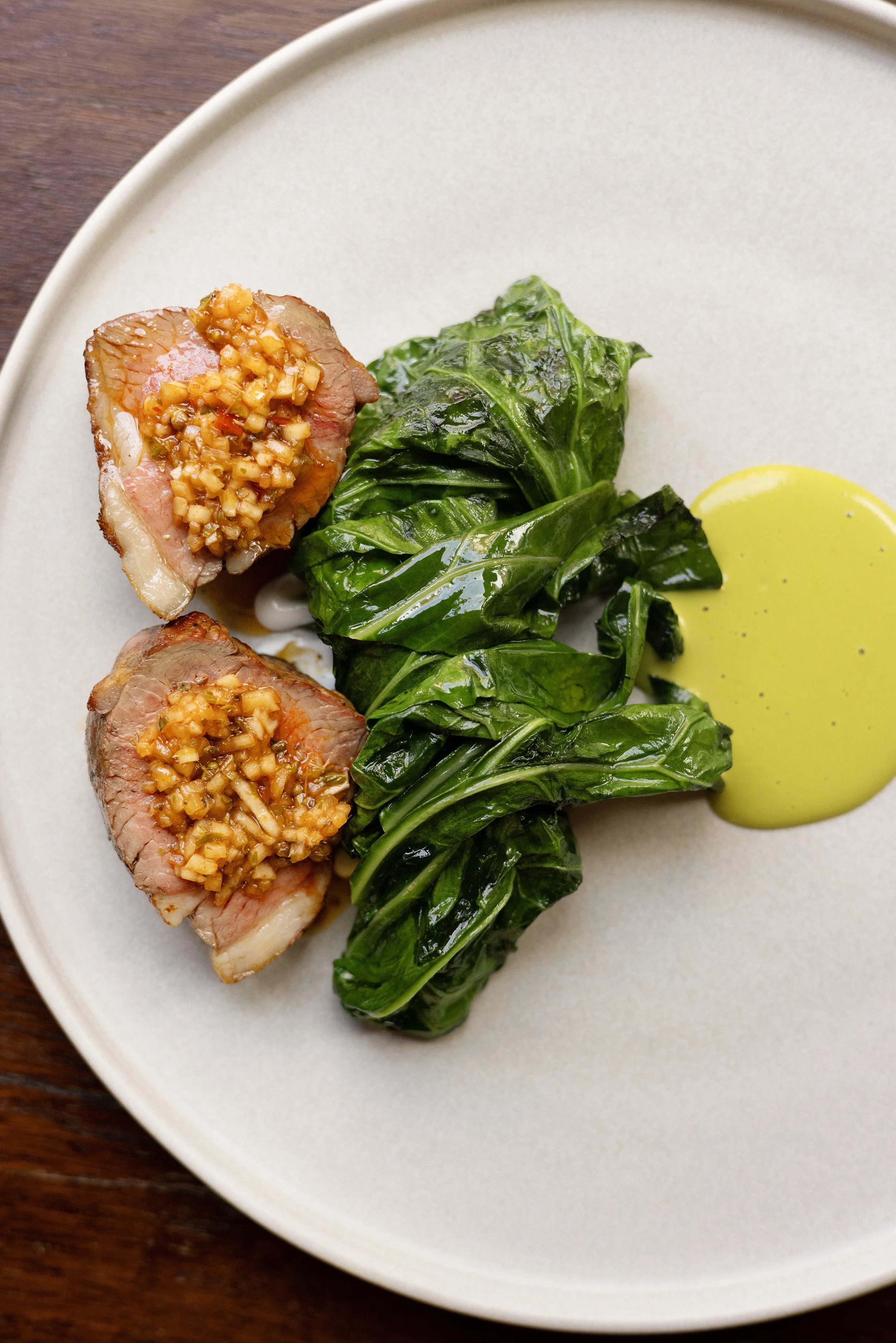 Two pieces of Lamb topped with a garlic and Pistachio sauce, served with sautéed greens and a dollop of mustard sauce on a white plate.