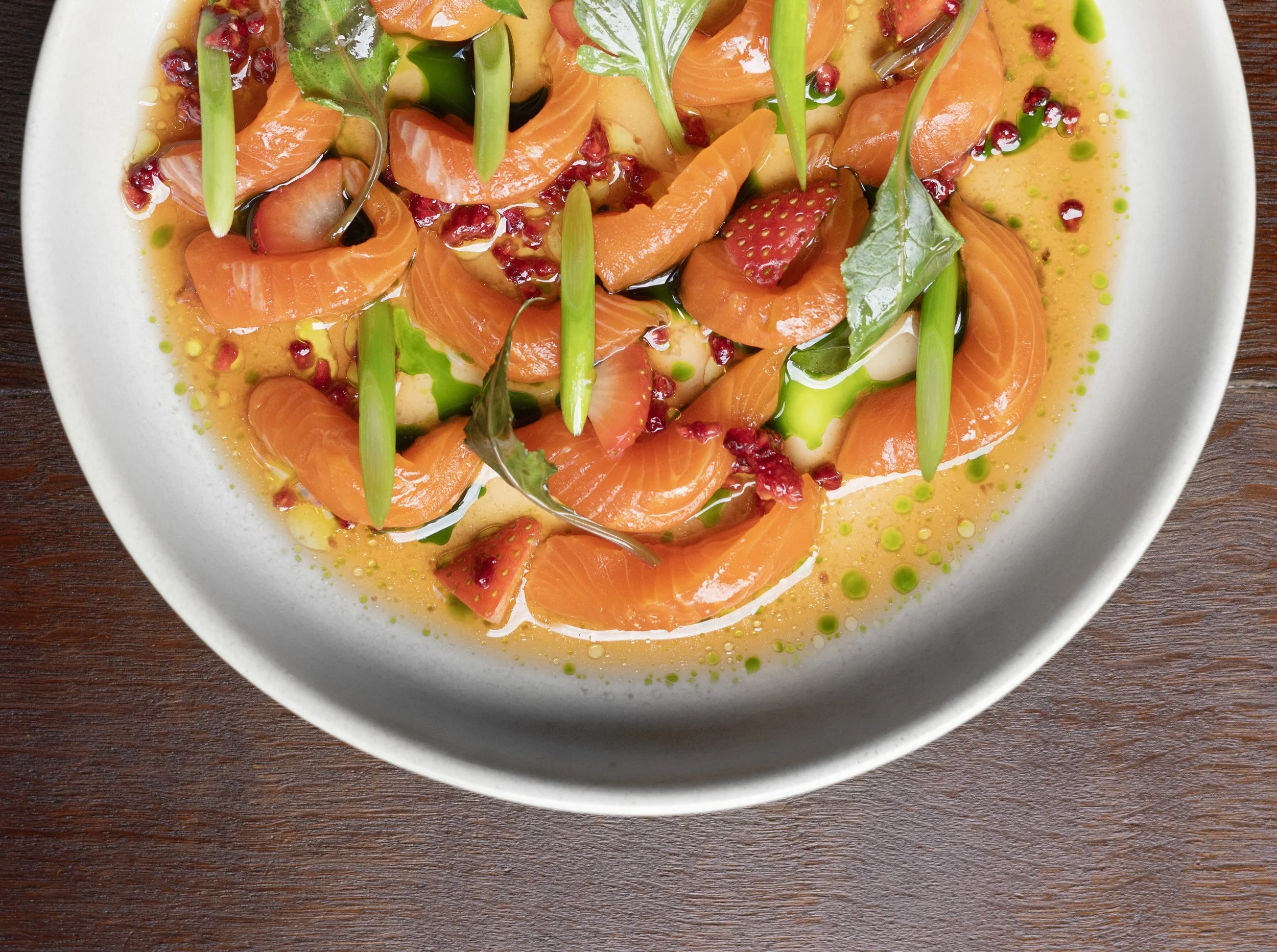 A white bowl of Trout sashimi with green onions, strawberries, pomegranate seeds, and a ponzu sauce. Green drops of oil or sauce are drizzled on top. The bowl is on a dark wooden table.