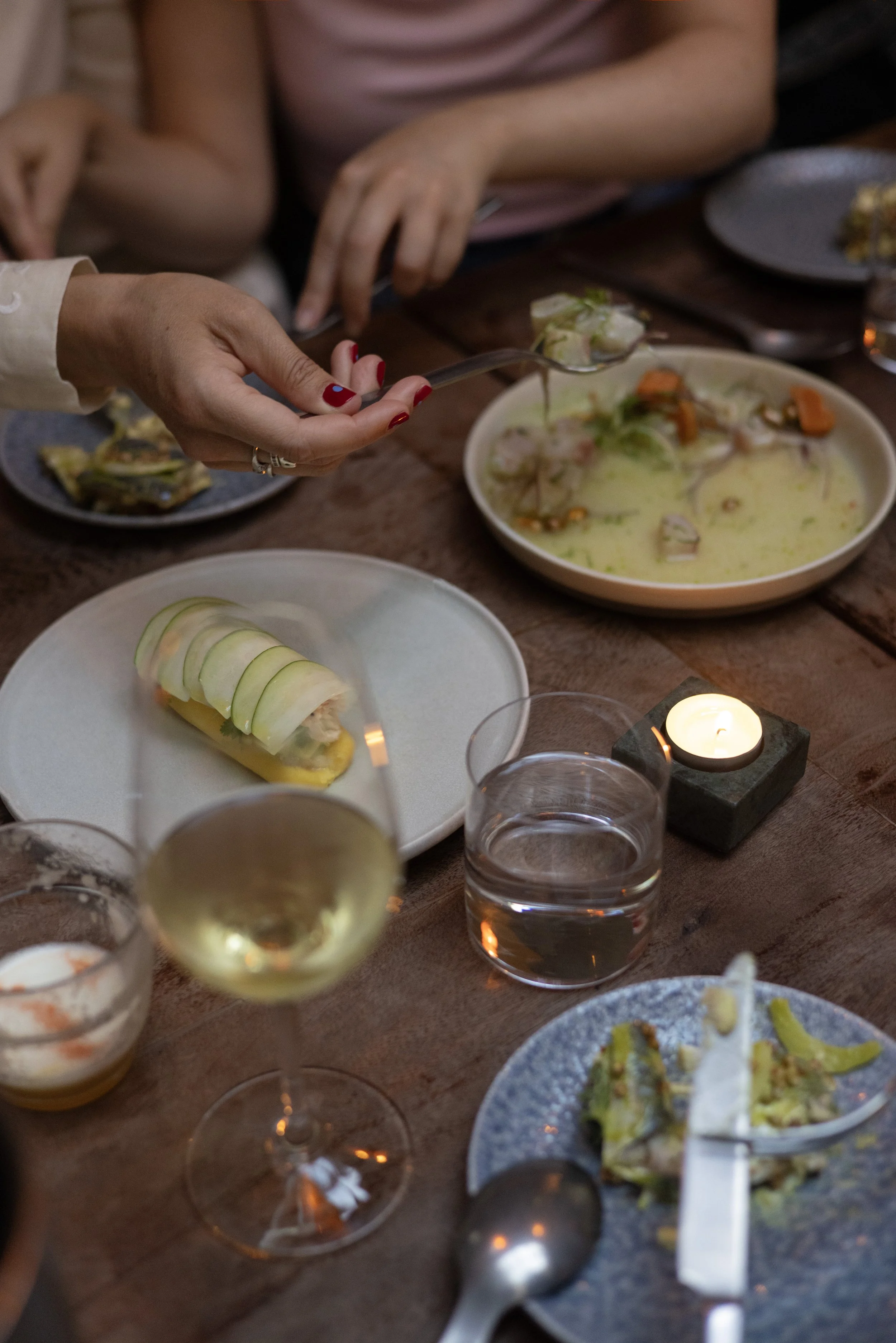 People sharing a meal with various dishes including Crab Causa, Ceviche, and finger foods, along with a glass of white wine and candles on a wooden table.
