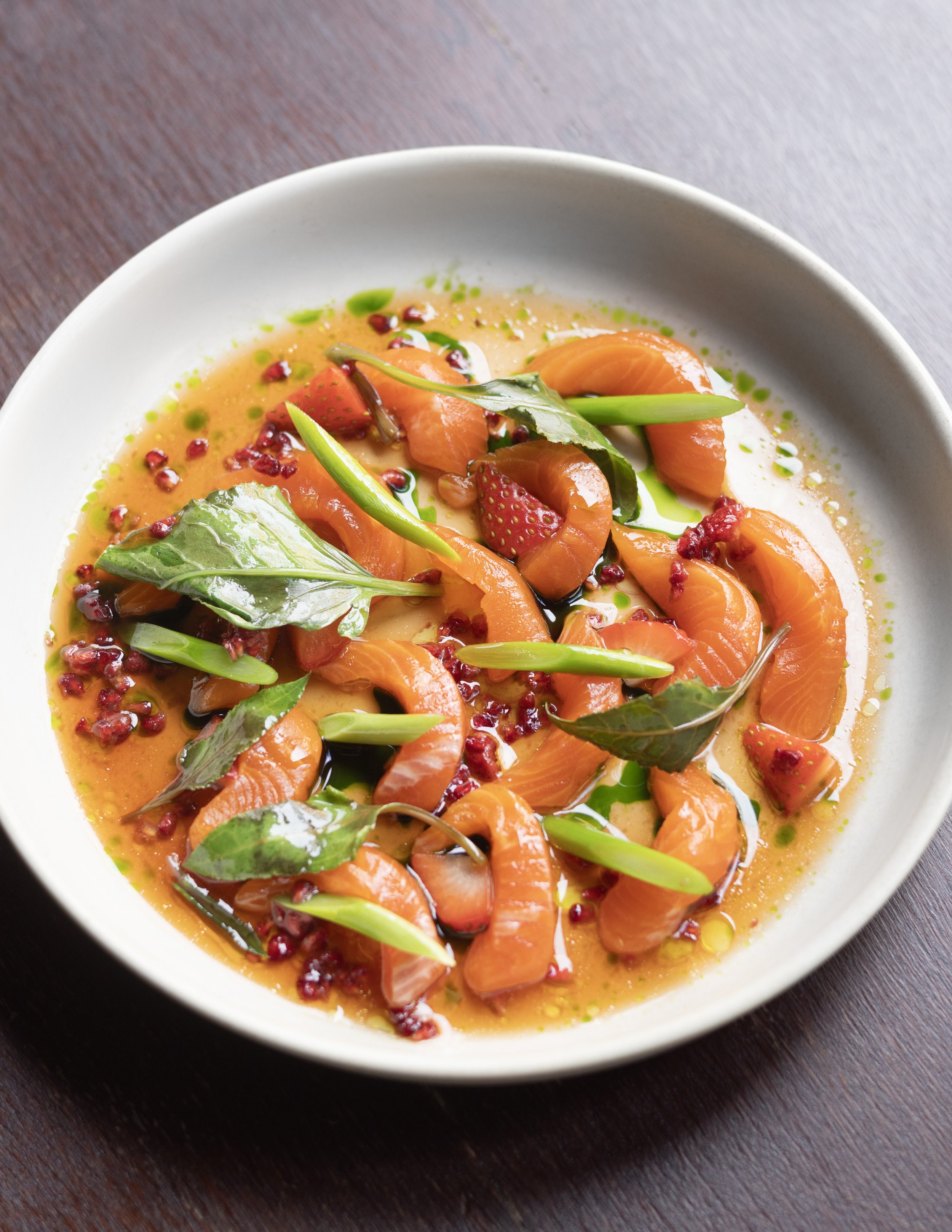 A bowl of trout with berries, green herbs, and a creamy sauce on a dark wooden table.