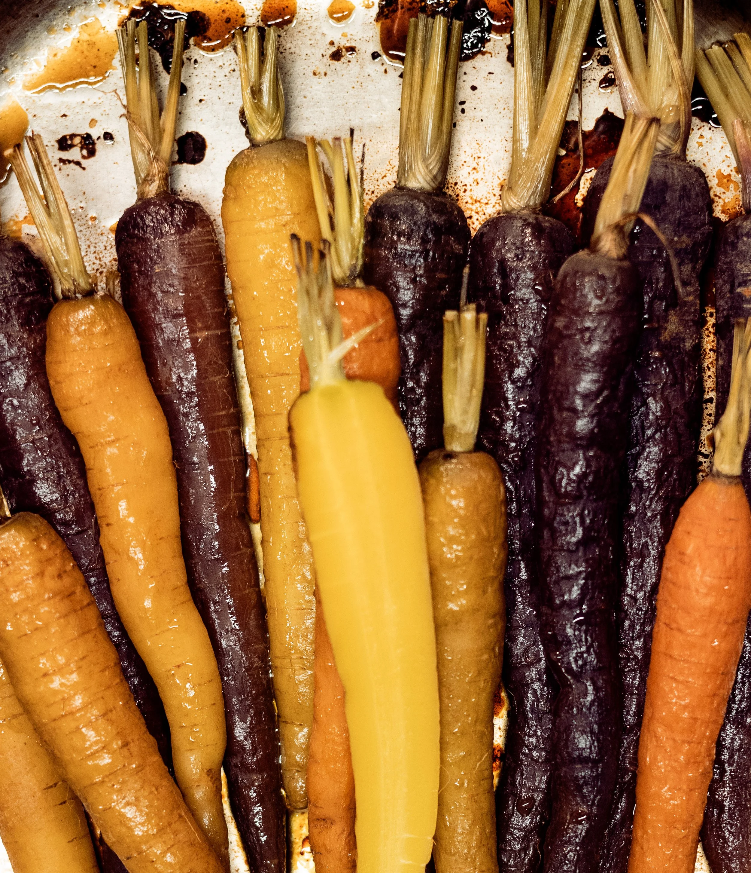 Colorful variety of carrots with green tops on a baking sheet, some with charred skin 