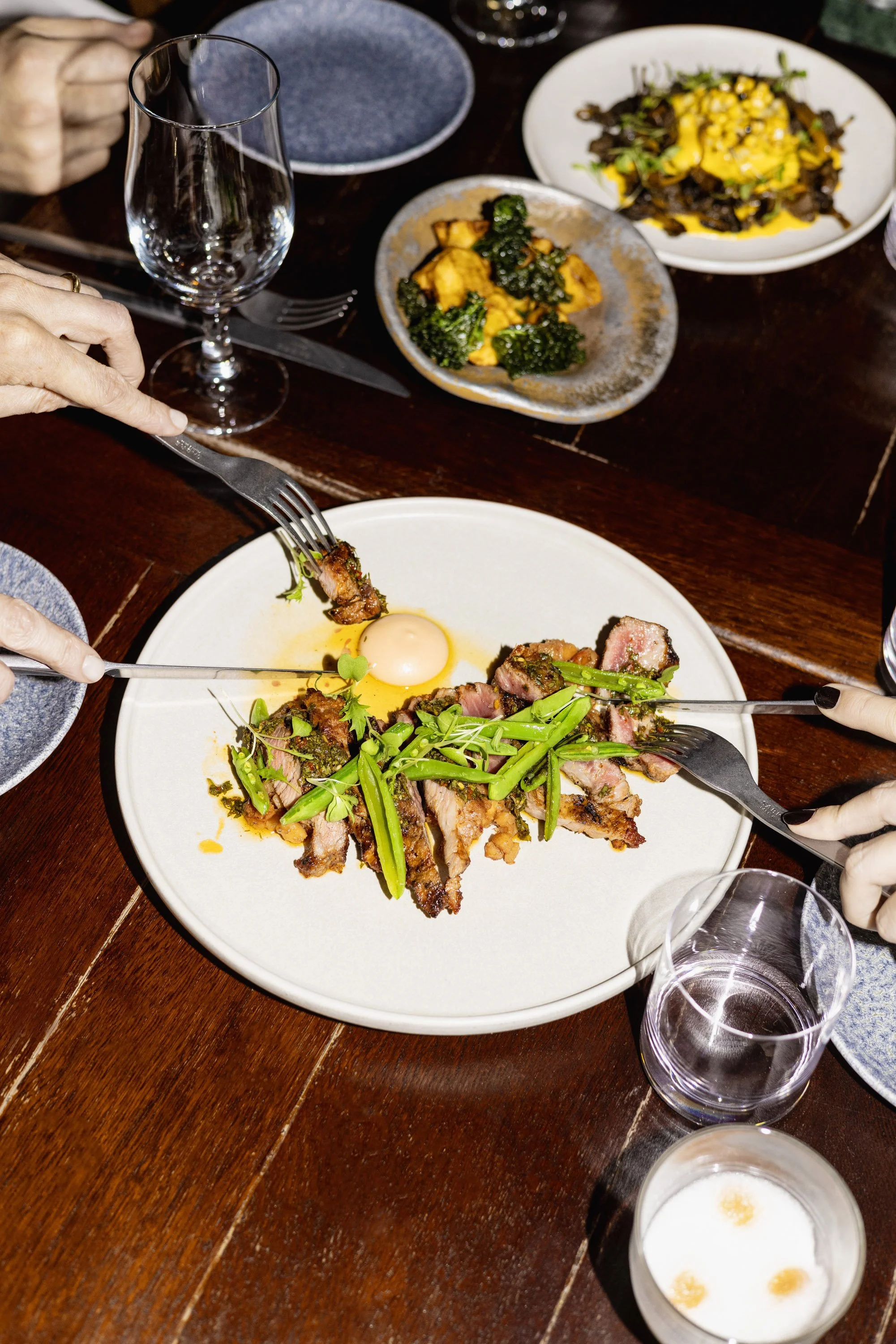 A dinner table with several plates of food, glasses of water, and hands using forks and knives to eat. There are grilled meat with green vegetables, a side of roasted potatoes, and a salad with a yellow dressing.
