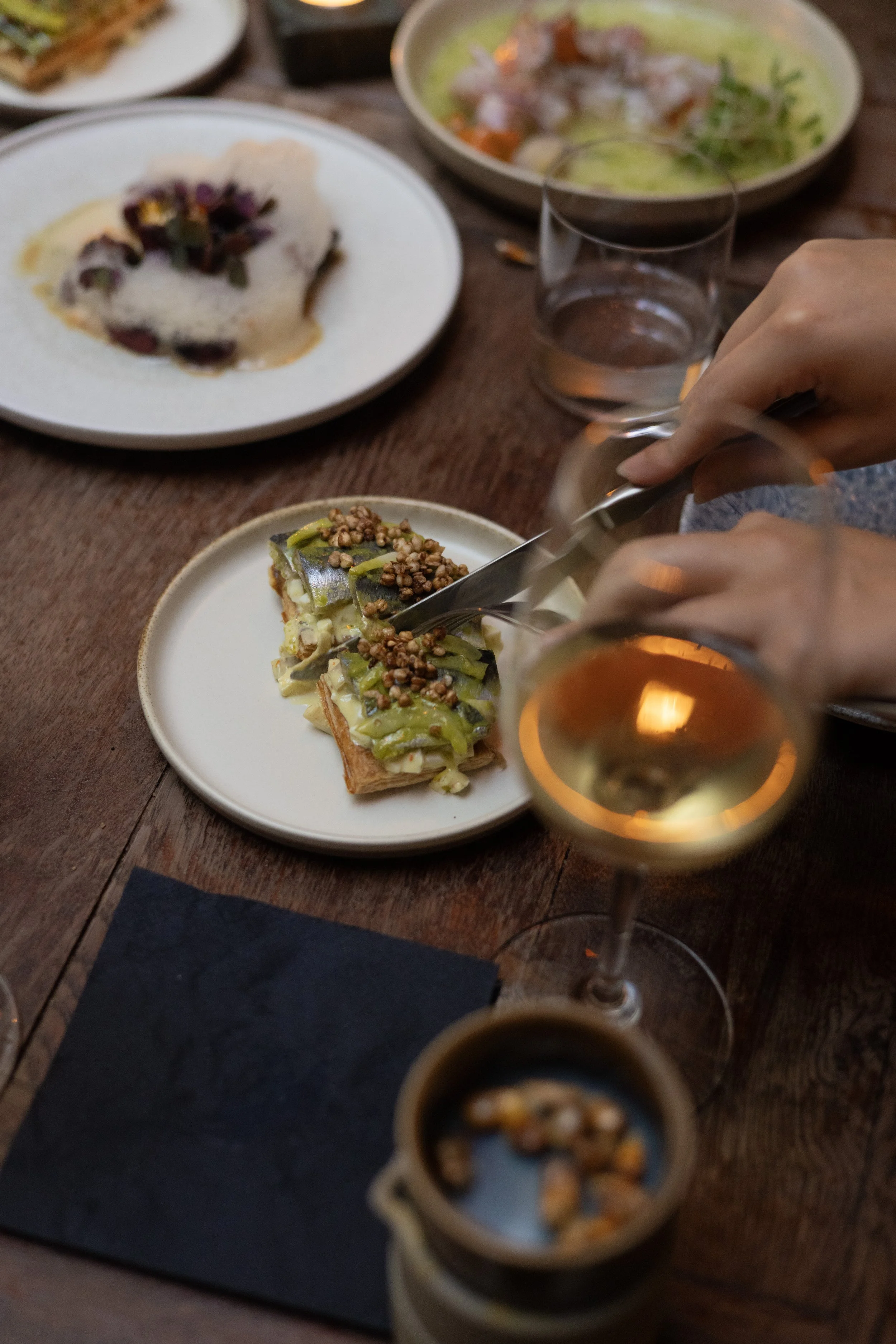 A dinner table with various dishes, including a piece of toast with Sardines, toppings and a Ceviche. There are also glasses of water and a glass of wine or champagne.