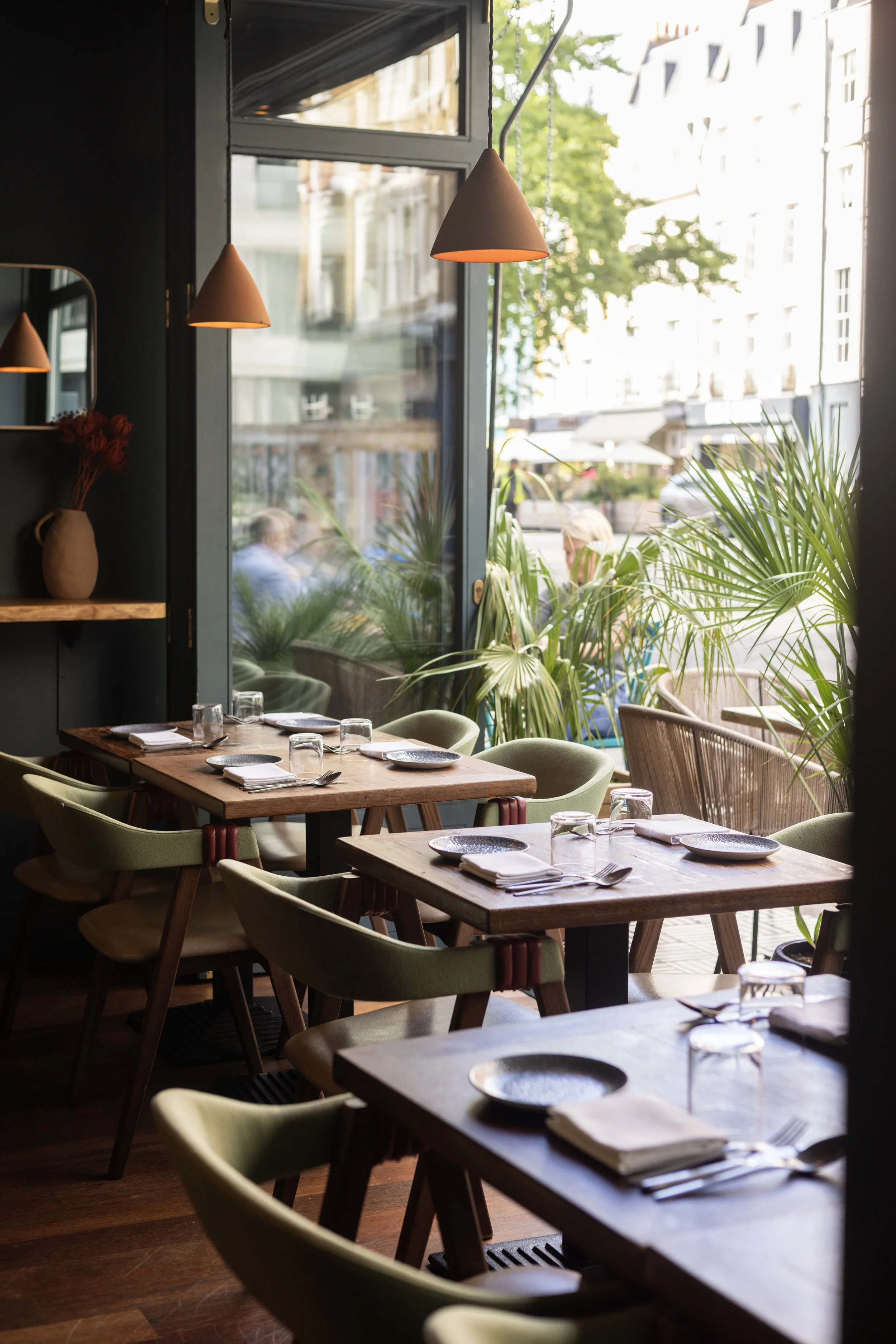 Interior of a LIMA FITZROVIA with wooden tables, green upholstered chairs, tropical plants, and large windows showing an outdoor street scene with buildings and pedestrians.