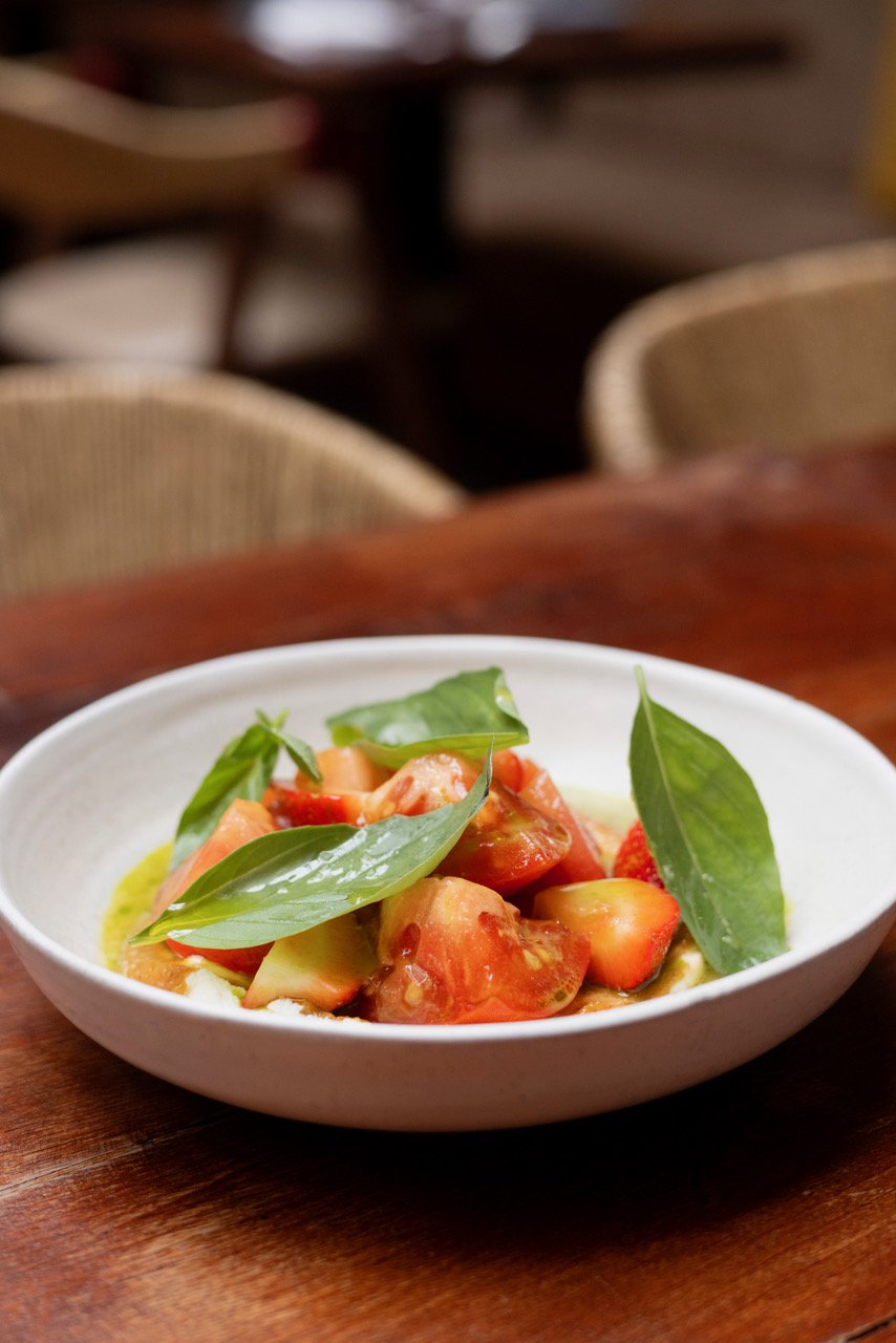 A white bowl with chopped tomatoes, fresh basil leaves, and olive oil on a wooden table.
