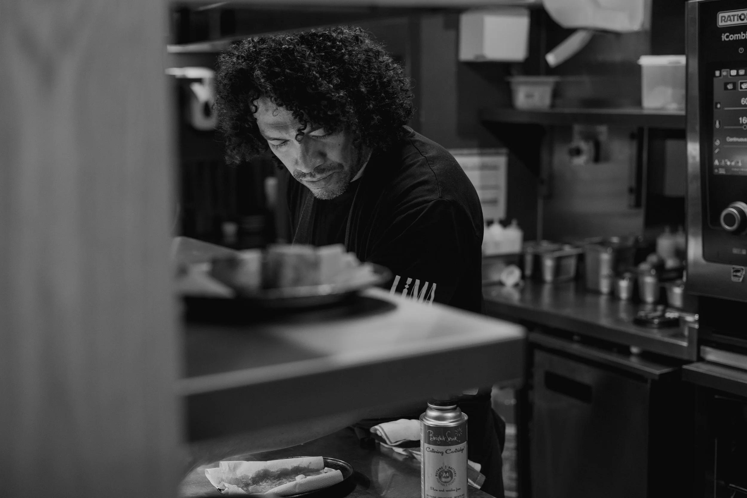 A man with curly hair looking down, working in a kitchen or food preparation area, with shelves, containers, and kitchen equipment in the background.