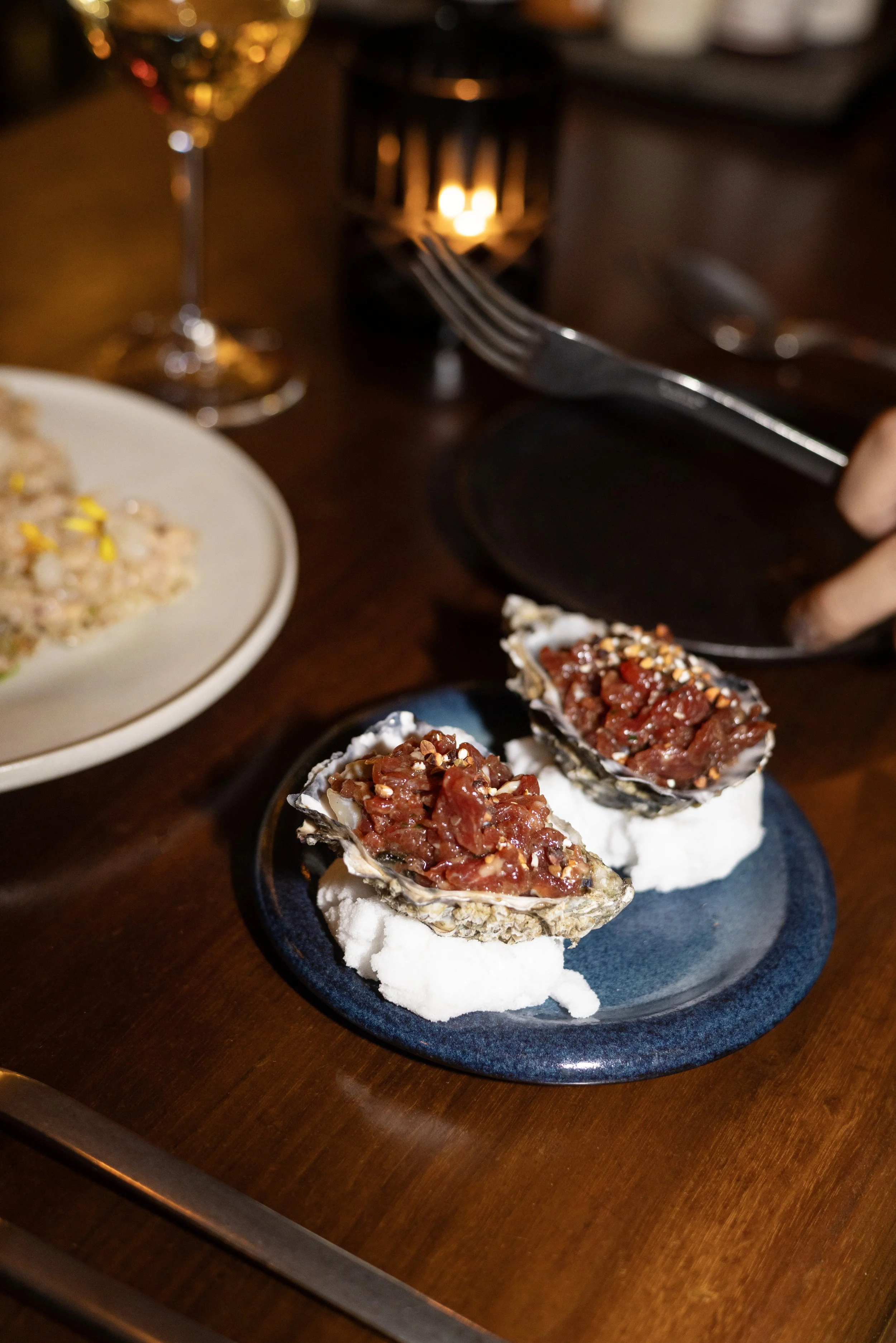 Close-up of two beef tatare & oysters topped with sesame seeds on a dark plate, with a glass of white wine, a fork, and some dishes in the blurry background.