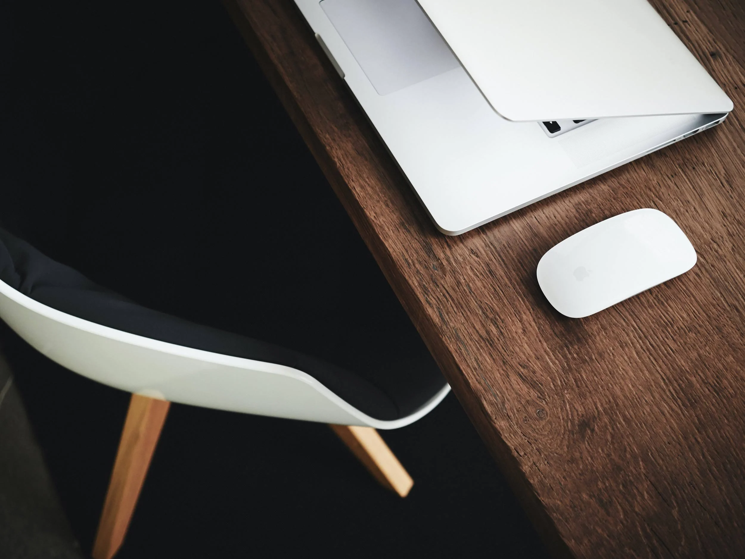 A silver laptop, a white wireless mouse, and a black and white chair at a wooden desk.