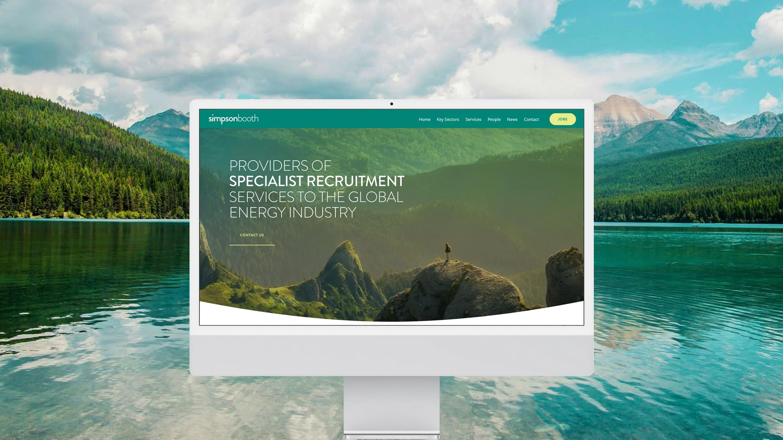 Computer monitor displaying a website for Simpson Booth, a provider of specialist recruitment services to the global energy industry, set outdoors with a lake, mountains, and forest in the background.