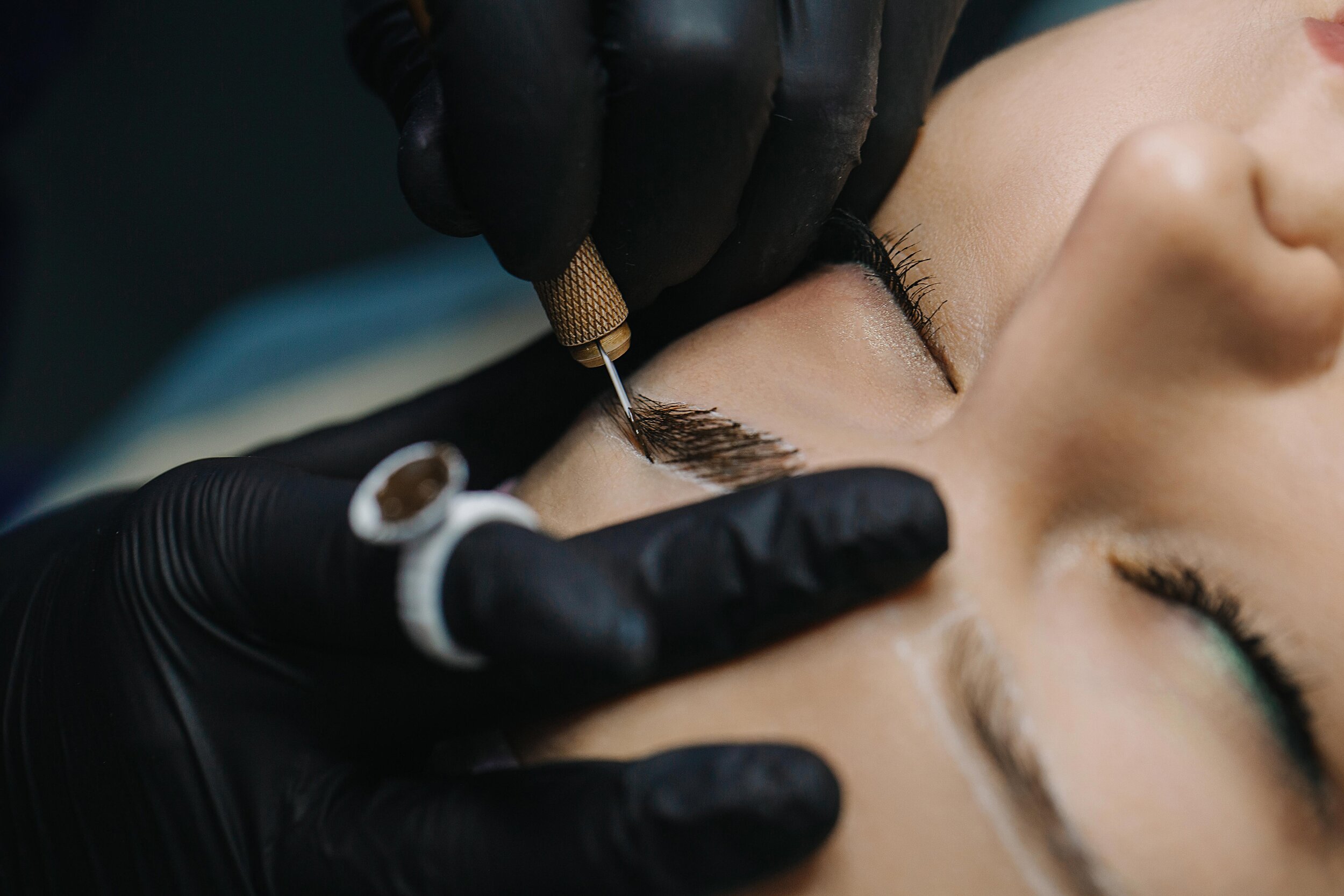 A person receiving a microblading procedure on their eyebrows, performed by a technician wearing black gloves.