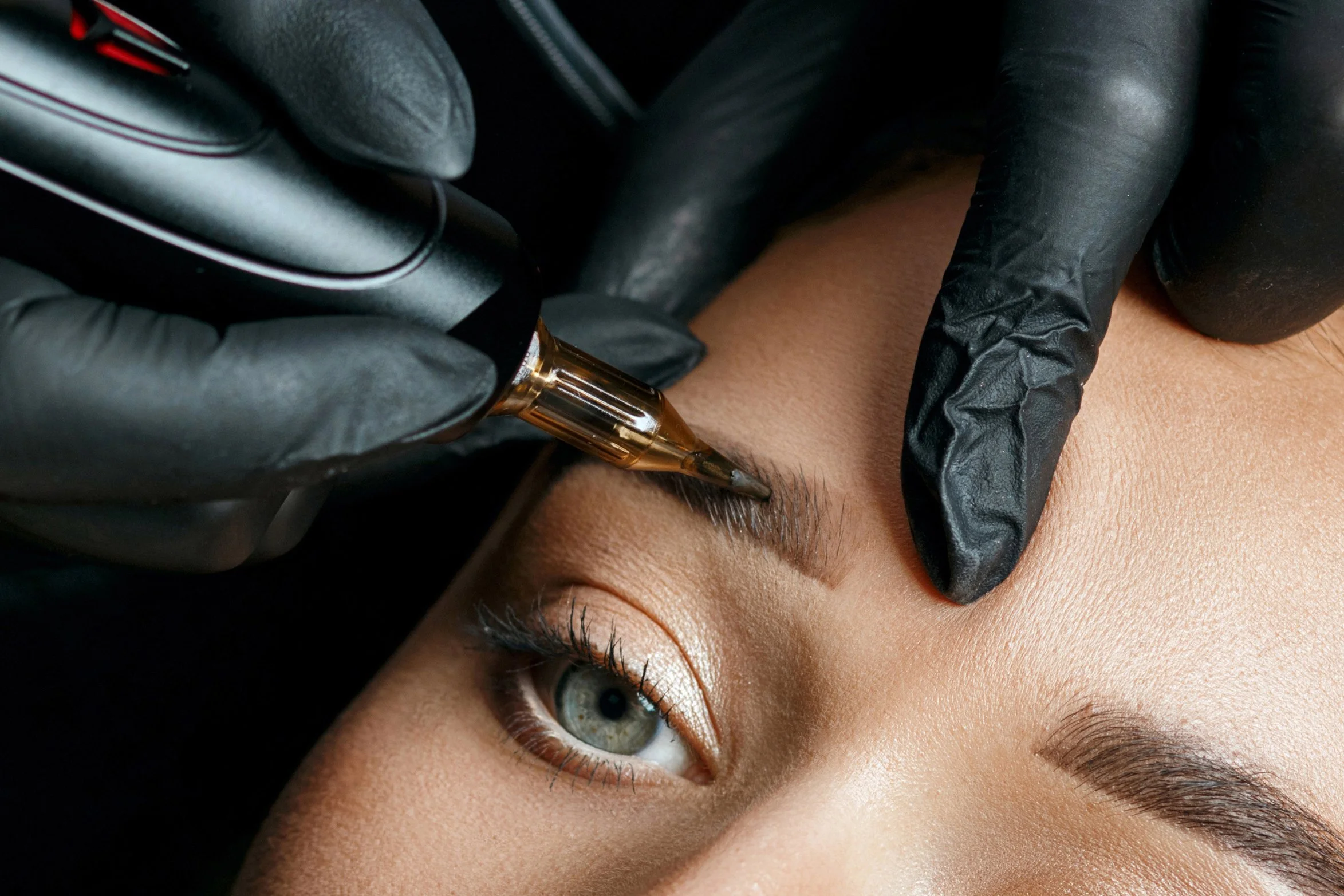 A person receiving an eyebrow tattooing or microblading procedure, with a tattoo artist wearing black gloves using a tattoo pen on the eyebrow.