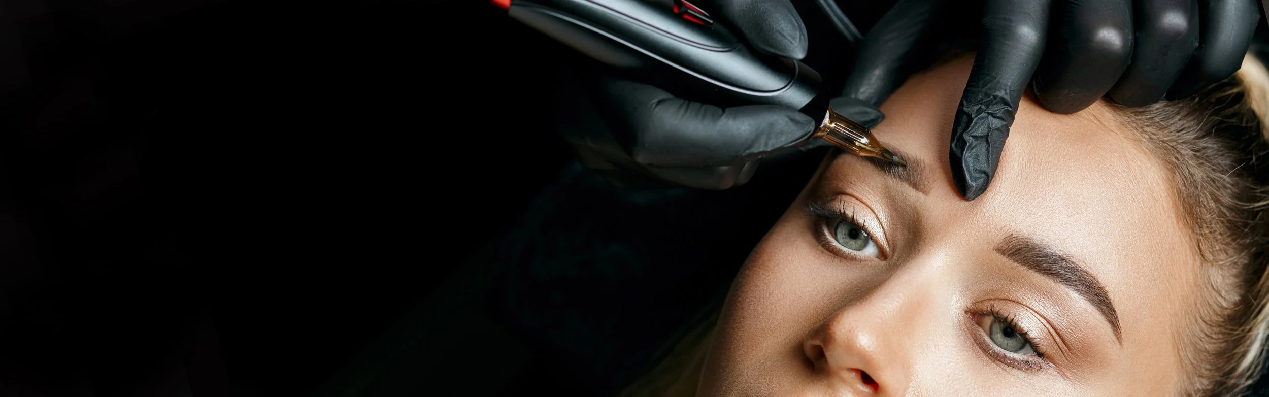 A woman receiving a tattoo inked on her eyebrow by a tattoo artist wearing black gloves.