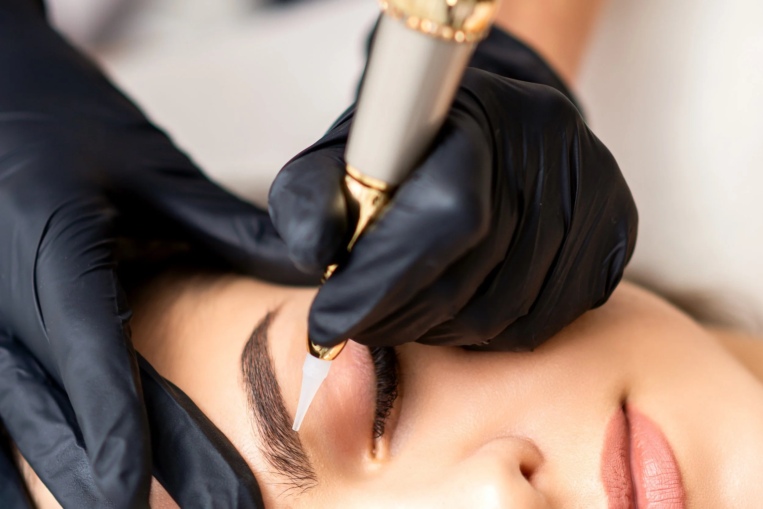 A person with tan skin getting a permanent makeup tattooing procedure on their eyebrows by a professional wearing black gloves, using a tattoo pen.