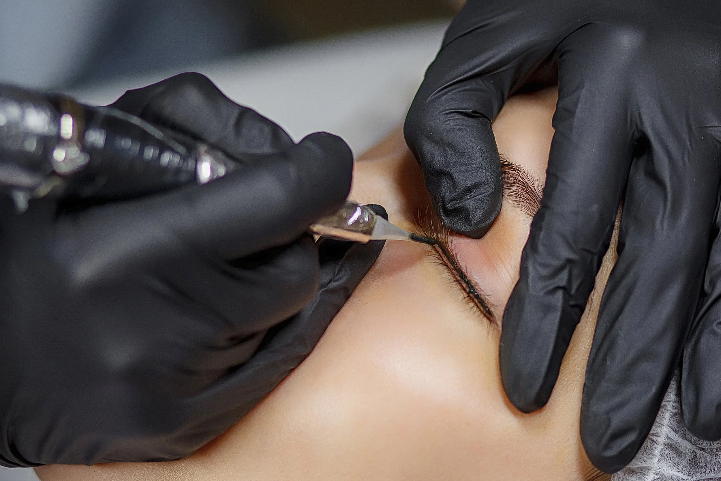 A person receiving a cosmetic tattooing procedure on their eyebrow using a tattoo machine, with individual wearing black gloves.