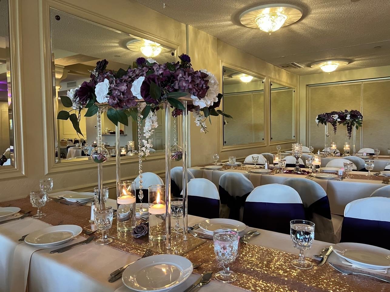 Elegant banquet hall set up for a formal event with round tables, floral centerpieces with purple and white flowers, candles, and arranged place settings with glassware and cutlery.