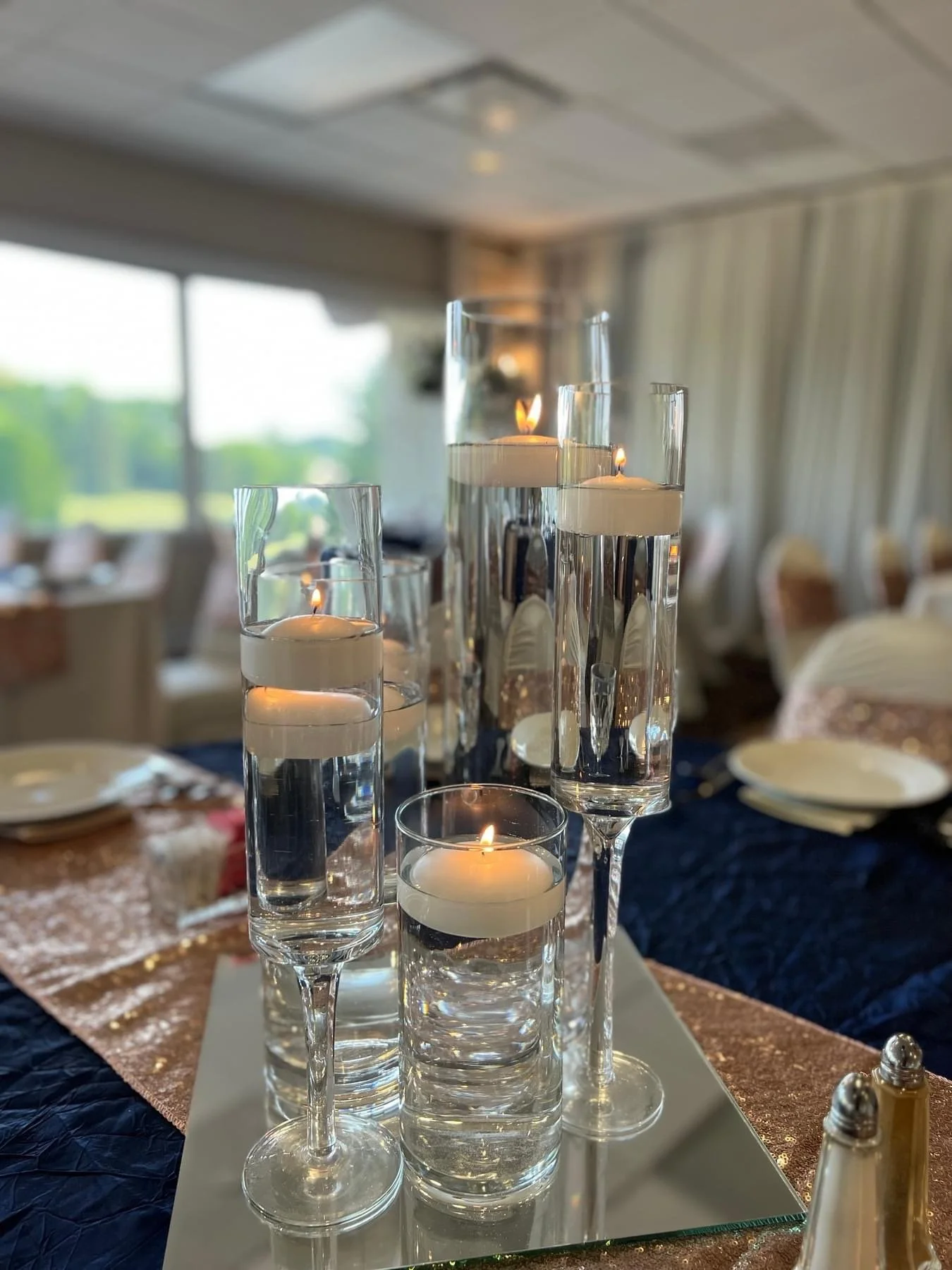 Table centerpiece with floating candles in glass vases of varying heights on a mirrored surface, set in a well-lit dining room with large windows and cream-colored curtains.