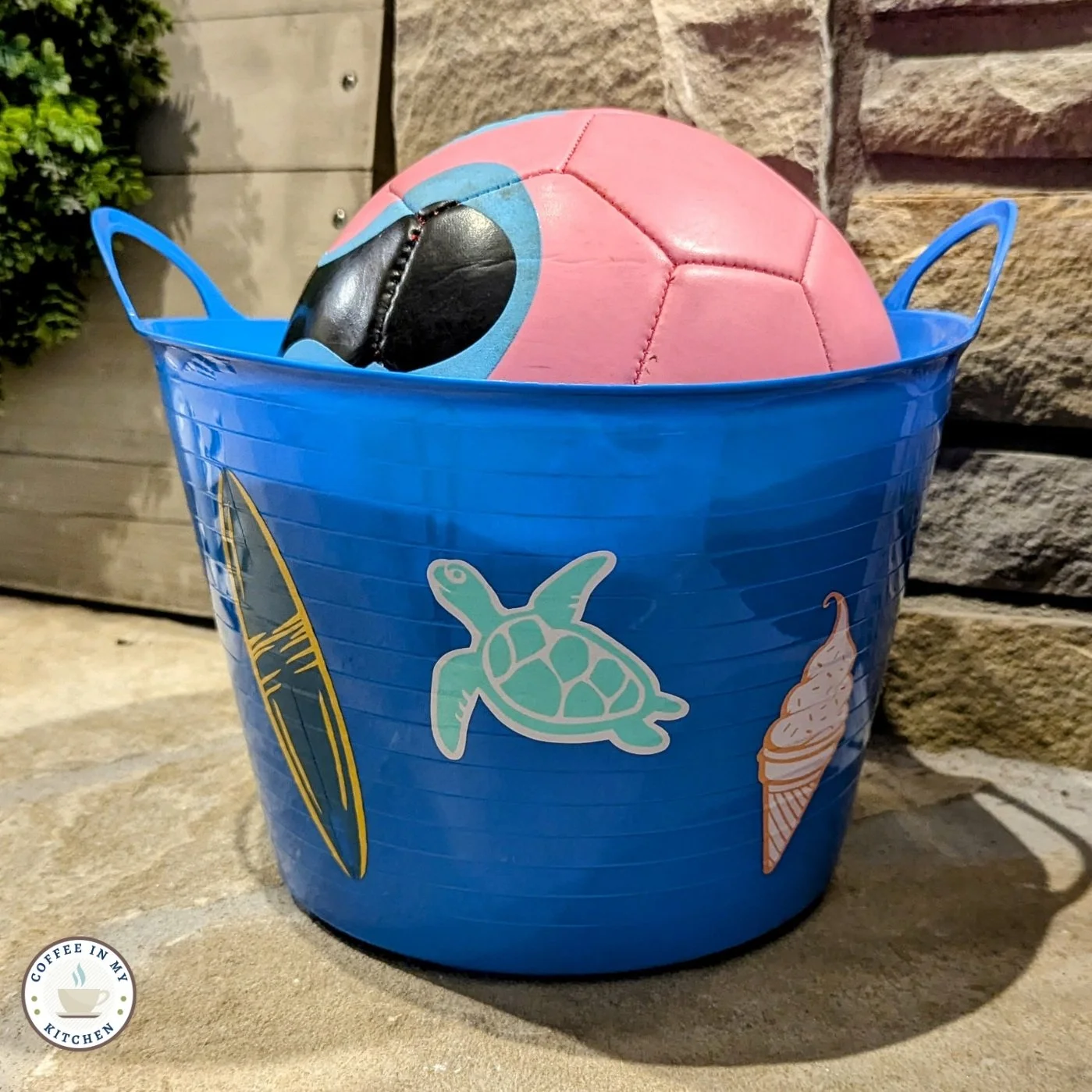 Blue bucket with a pink soccer ball, decorated with stickers of a surfboard, sea turtle, and ice cream cone, placed on a stone surface.