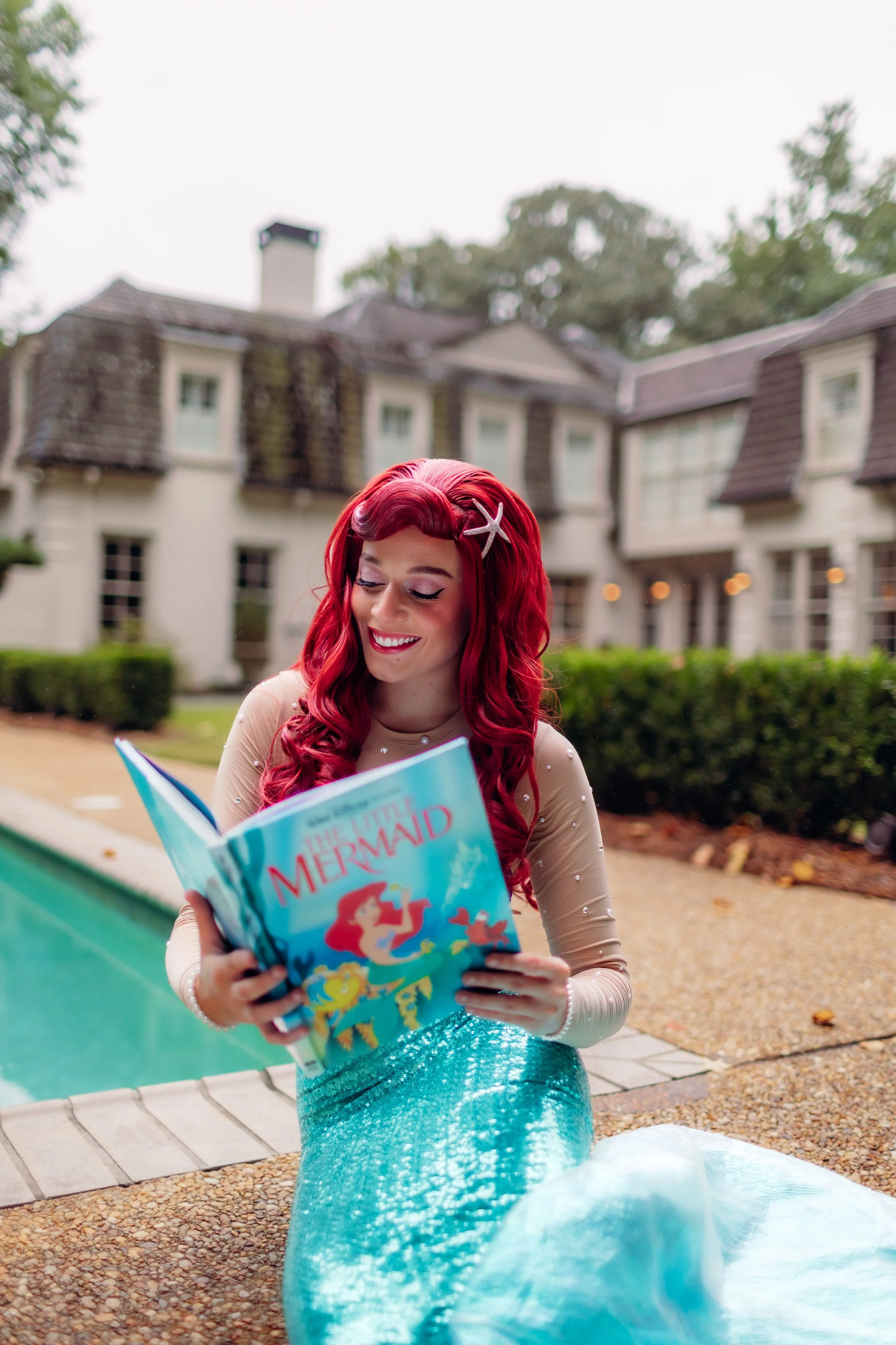 A woman dressed as a mermaid with red hair and a starfish hair accessory sitting by a pool, reading a book titled "The Little Mermaid" in front of a large house with a courtyard.