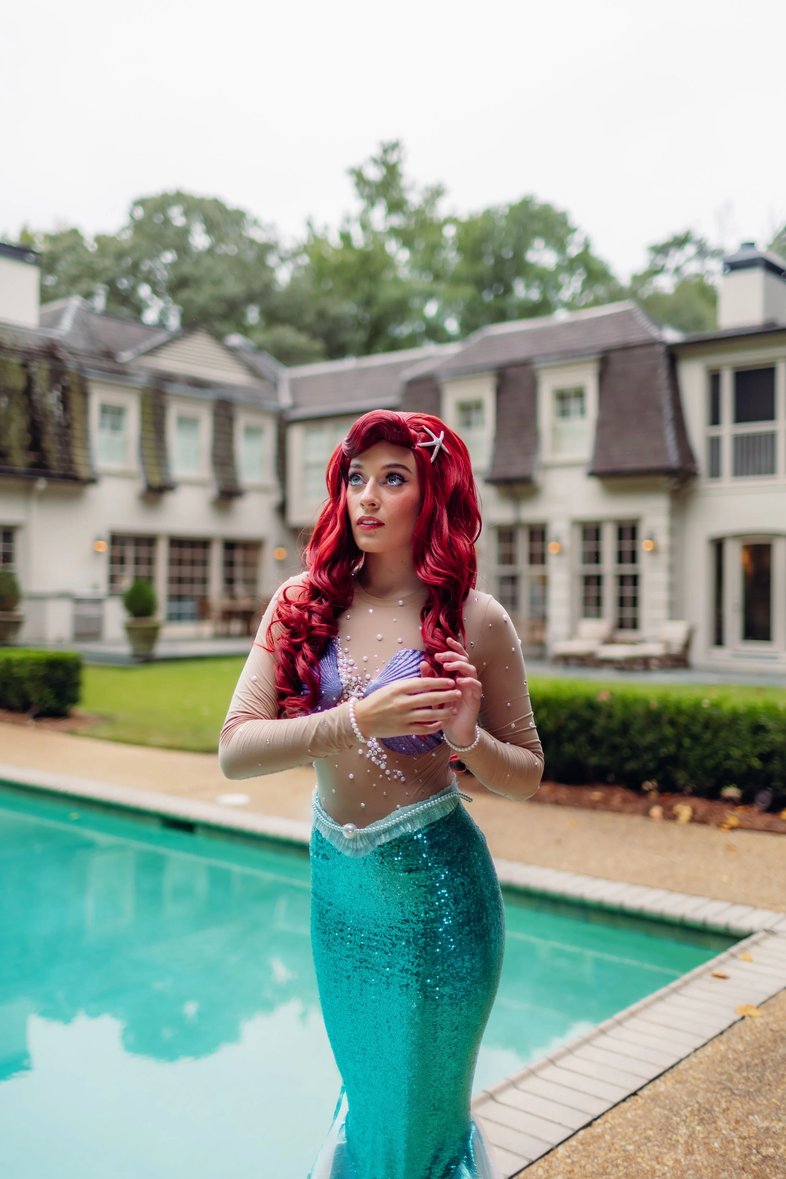A woman dressed as Ariel from Disney's The Little Mermaid standing outdoors near a swimming pool with a house in the background.