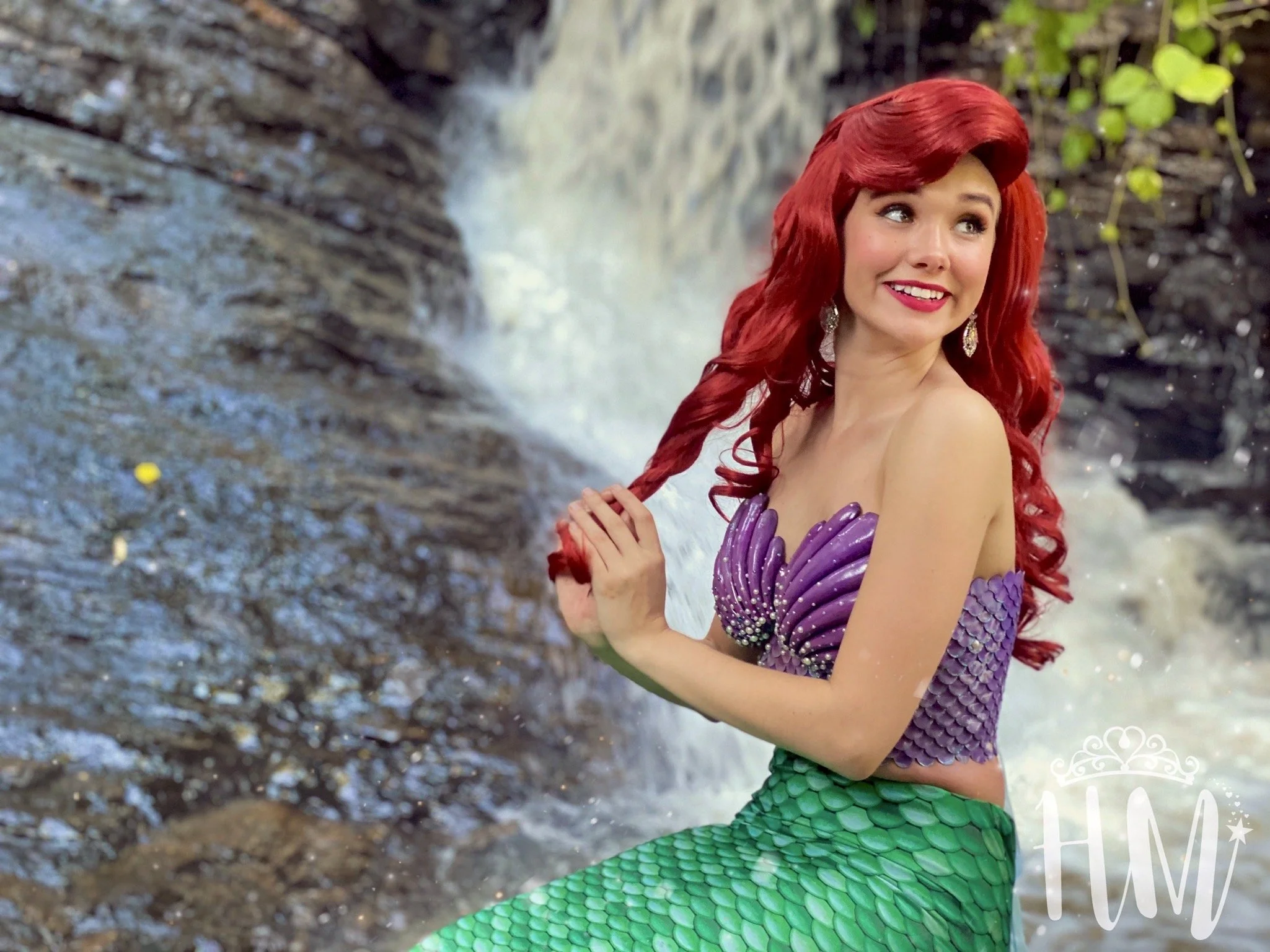 A woman dressed as a mermaid with long red hair, a purple shell top, and green scale tail, sitting by a waterfall in a natural setting.