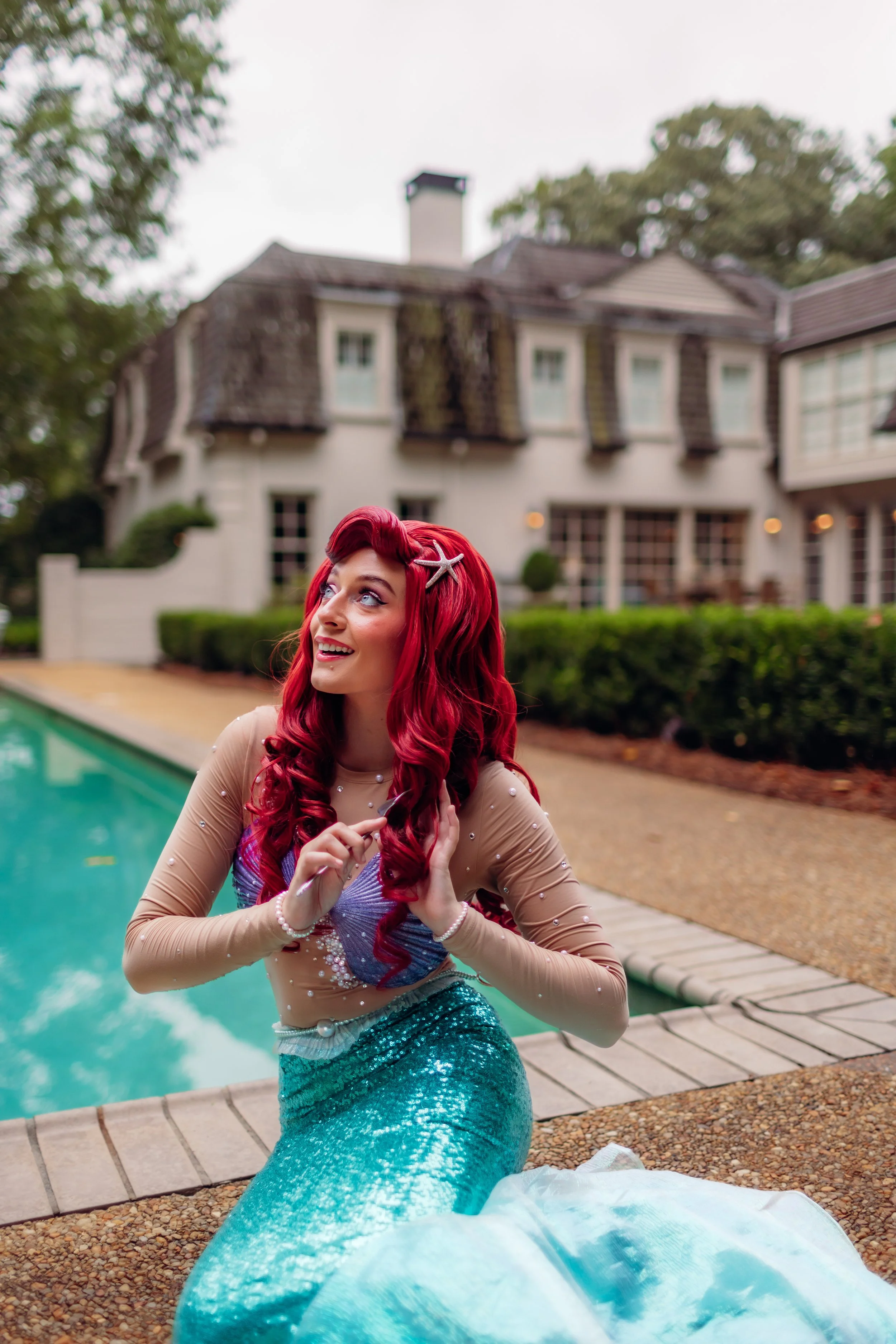 A woman in a mermaid costume sitting by a swimming pool in front of a large house with a tiled roof and multiple windows.